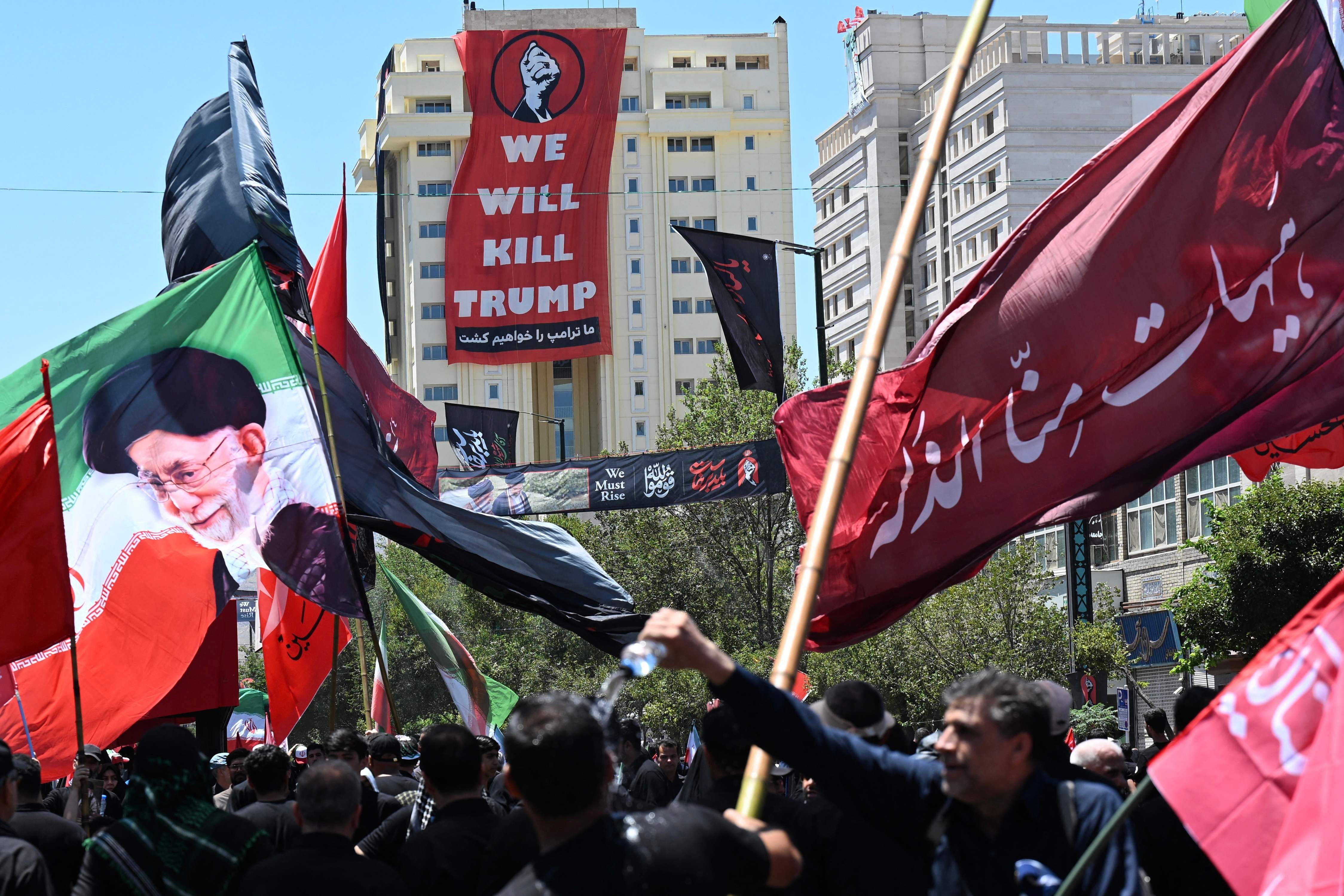 Menschenmenge mit Fahnen bei einer Trauerprozession; Banner mit „We will kill Trump“ im Hintergrund.
