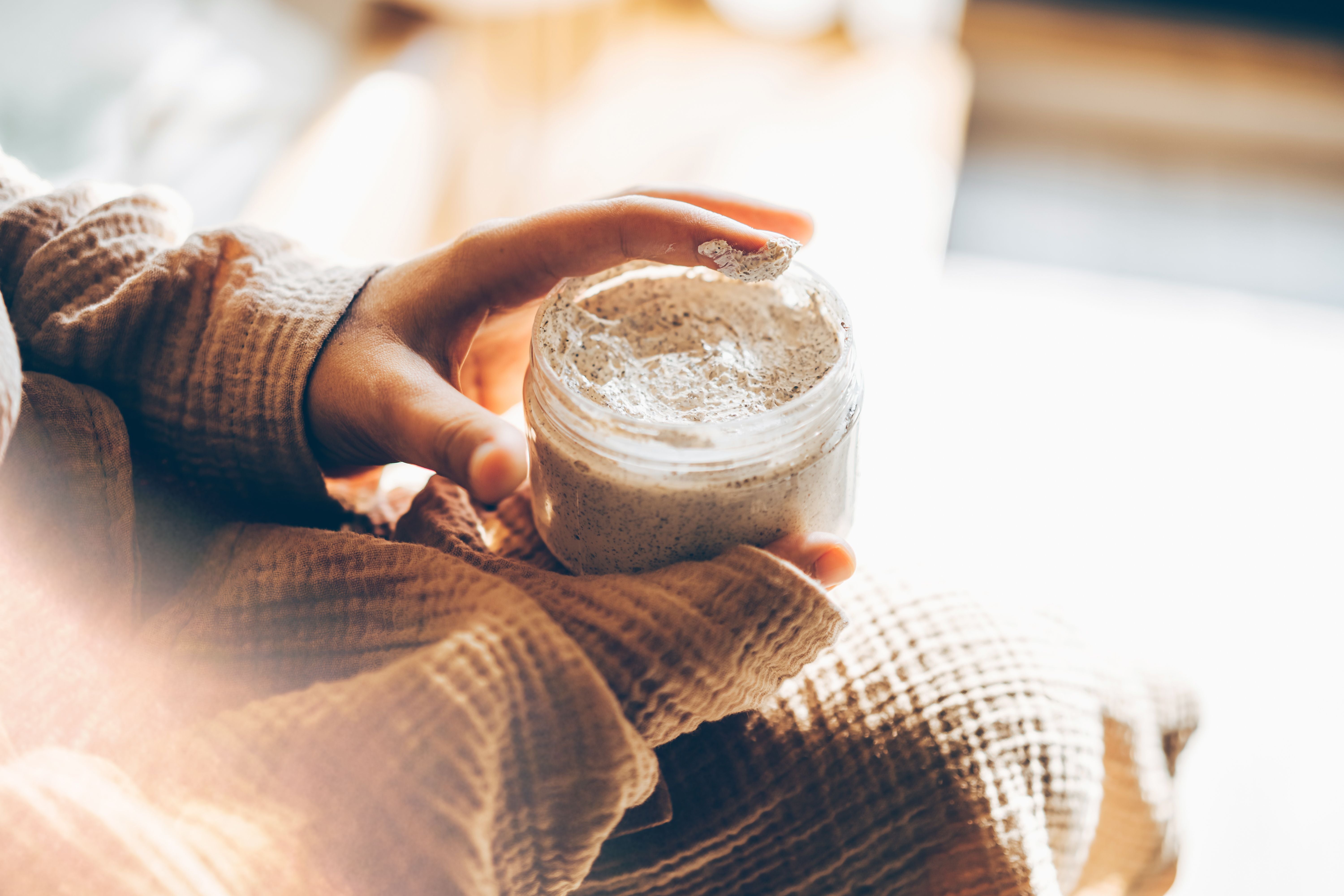Hand hält ein Glas mit Sheabutter-Peeling, Finger entnehmen etwas, warmes Sonnenlicht fällt darauf.