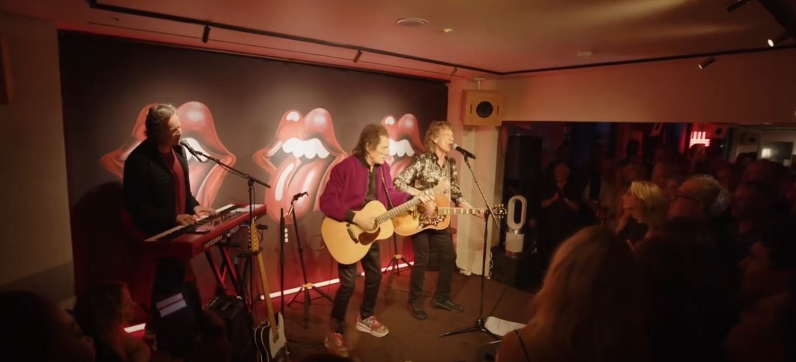 Drei Musiker der Rolling Stones spielen live vor einem Publikum, im Hintergrund das Zungen-Logo.