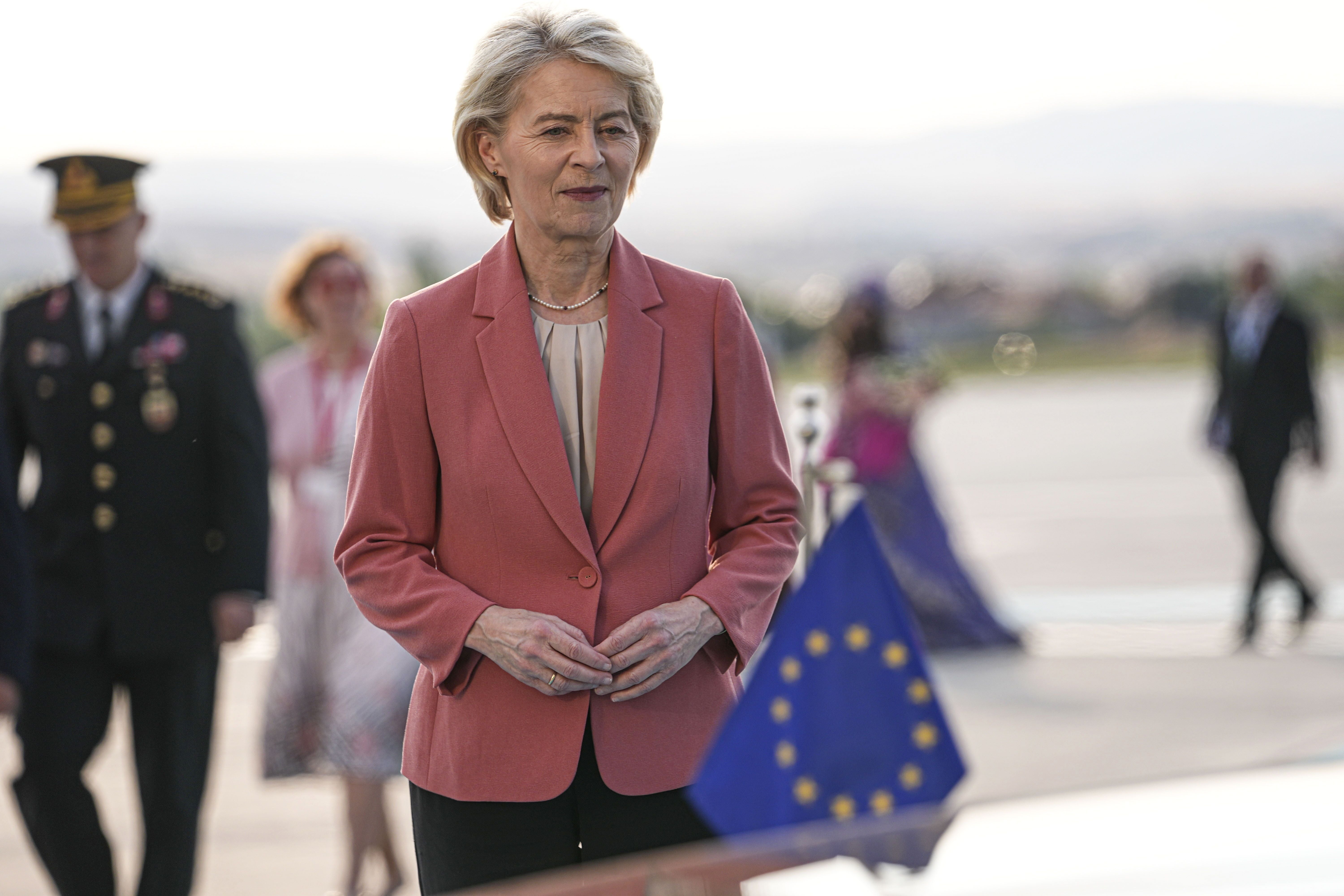 Ursula von der Leyen am Flughafen mit EU-Flagge im Vordergrund, Menschen im Hintergrund.