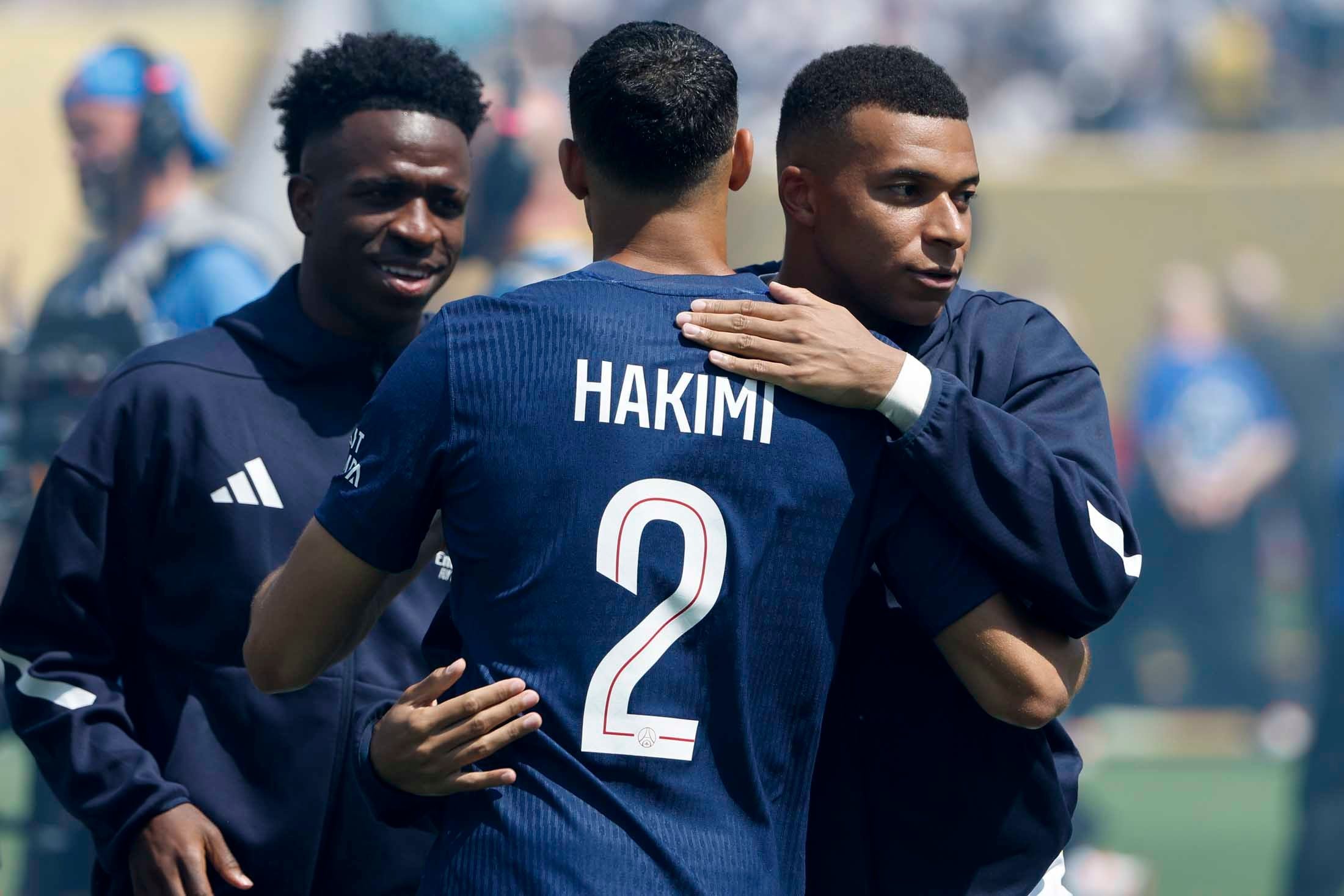 Ein Fußballer von hinten im dunkelblauen Trikot mit der Aufschrift HAKIMI und der Nummer 2 wird von einem anderen Spieler umarmt. Links im Hintergrund steht ein lächelnder Mann im Trainingsanzug.
