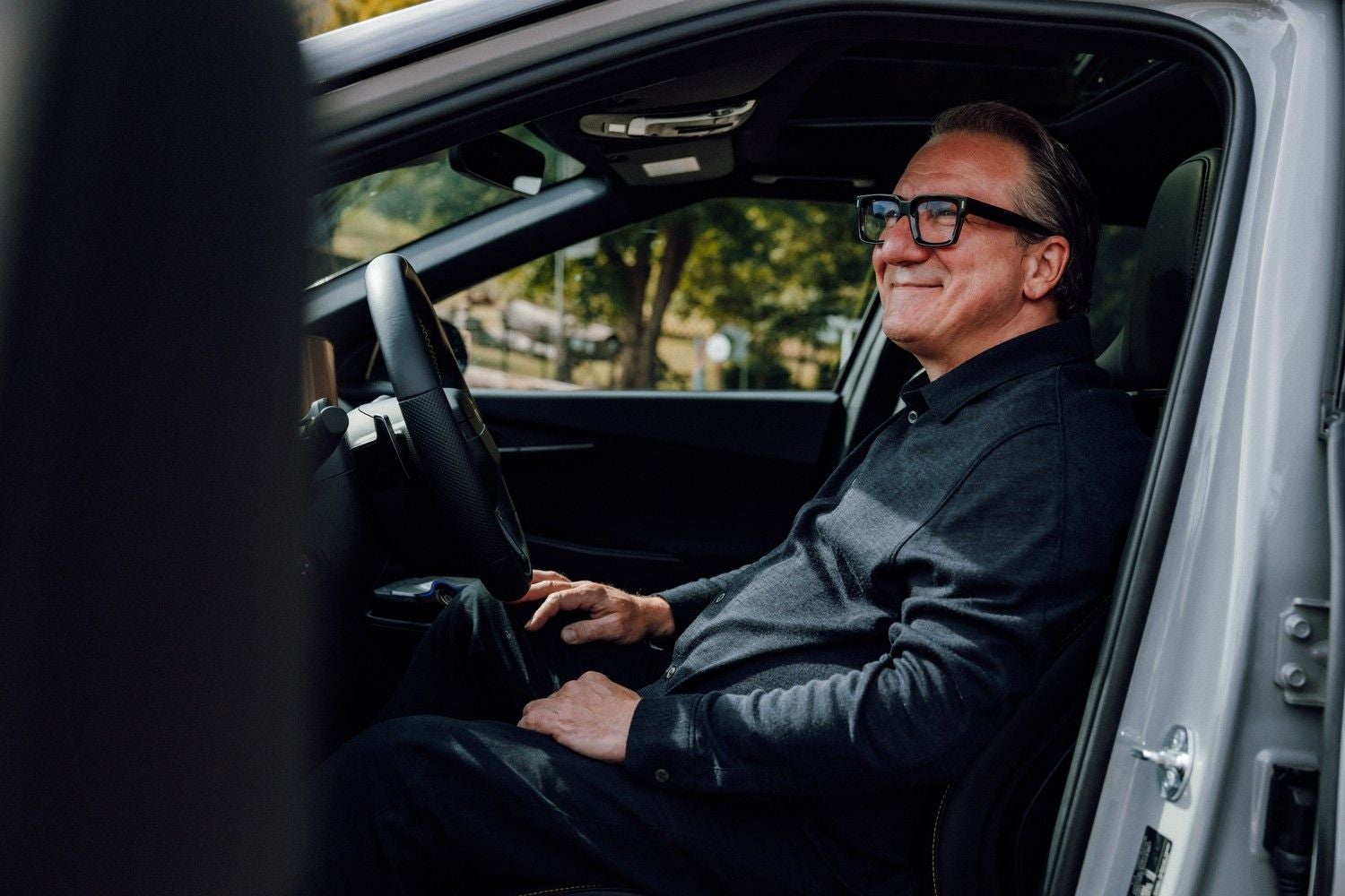 Ein älterer Mann mit Brille sitzt lächelnd am Steuer eines Autos.