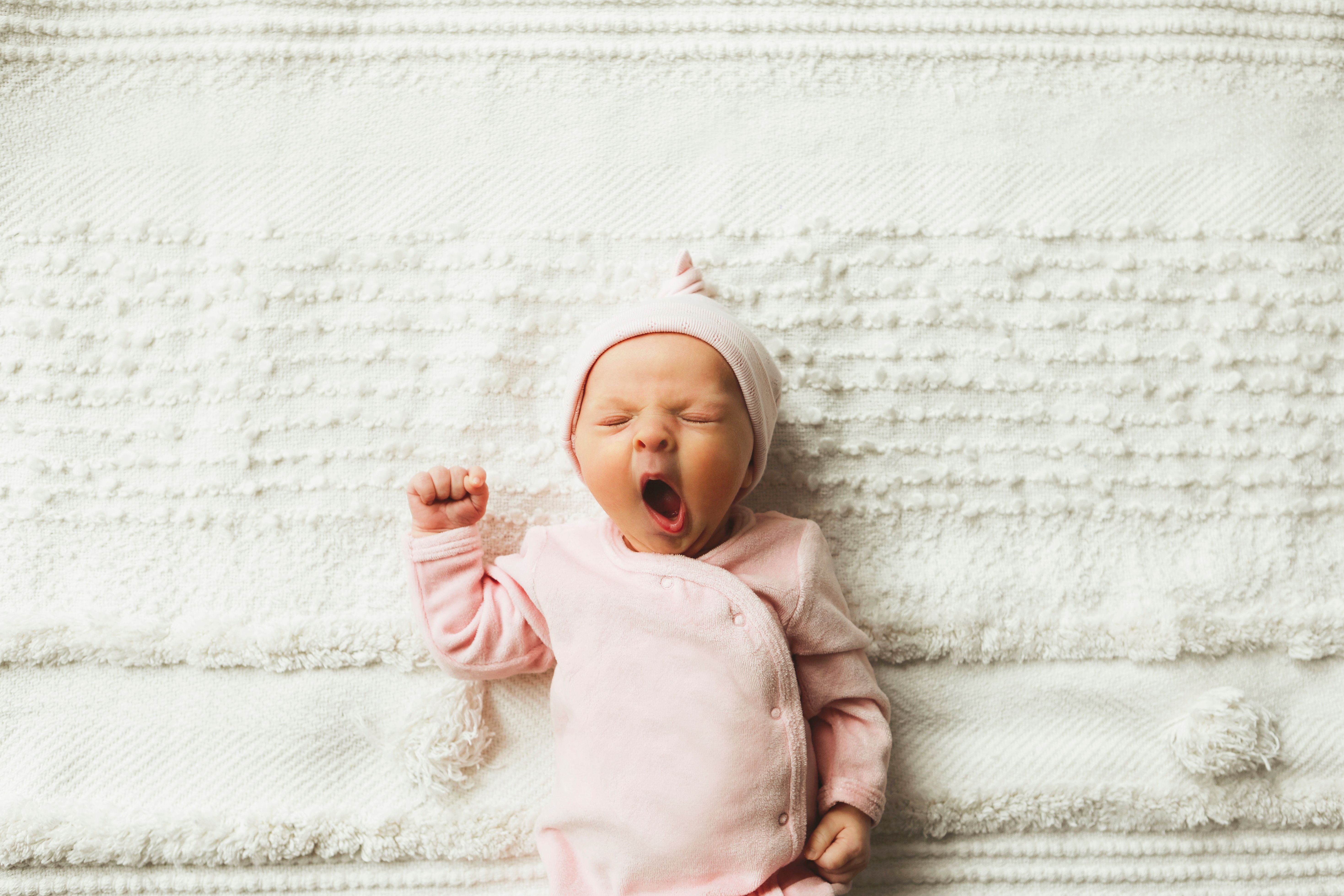 Gähnendes Baby in rosa Kleidung liegt auf einer weißen Decke.
