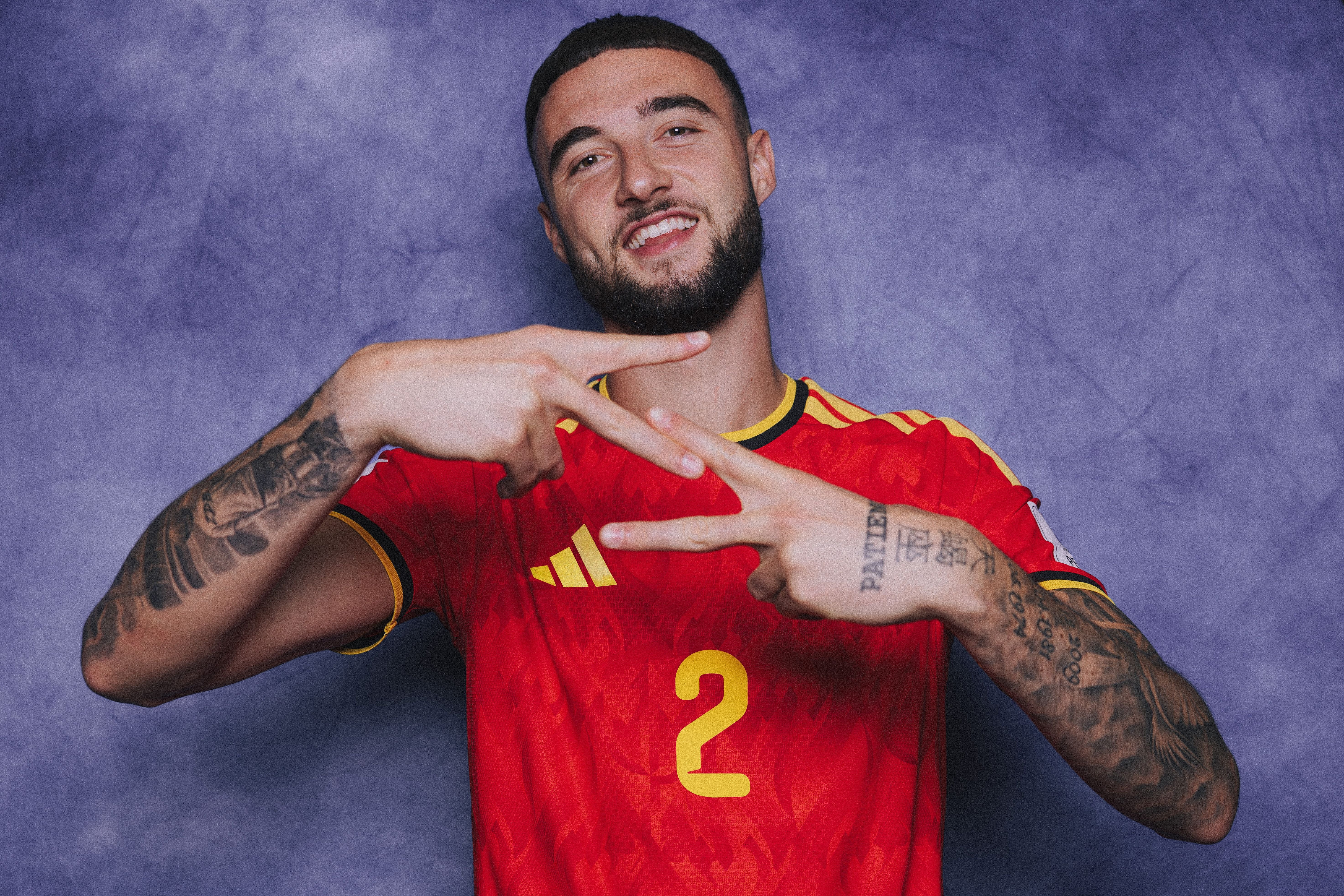 Ein Fußballspieler im roten Belgien-Trikot posiert lächelnd vor lila Hintergrund.