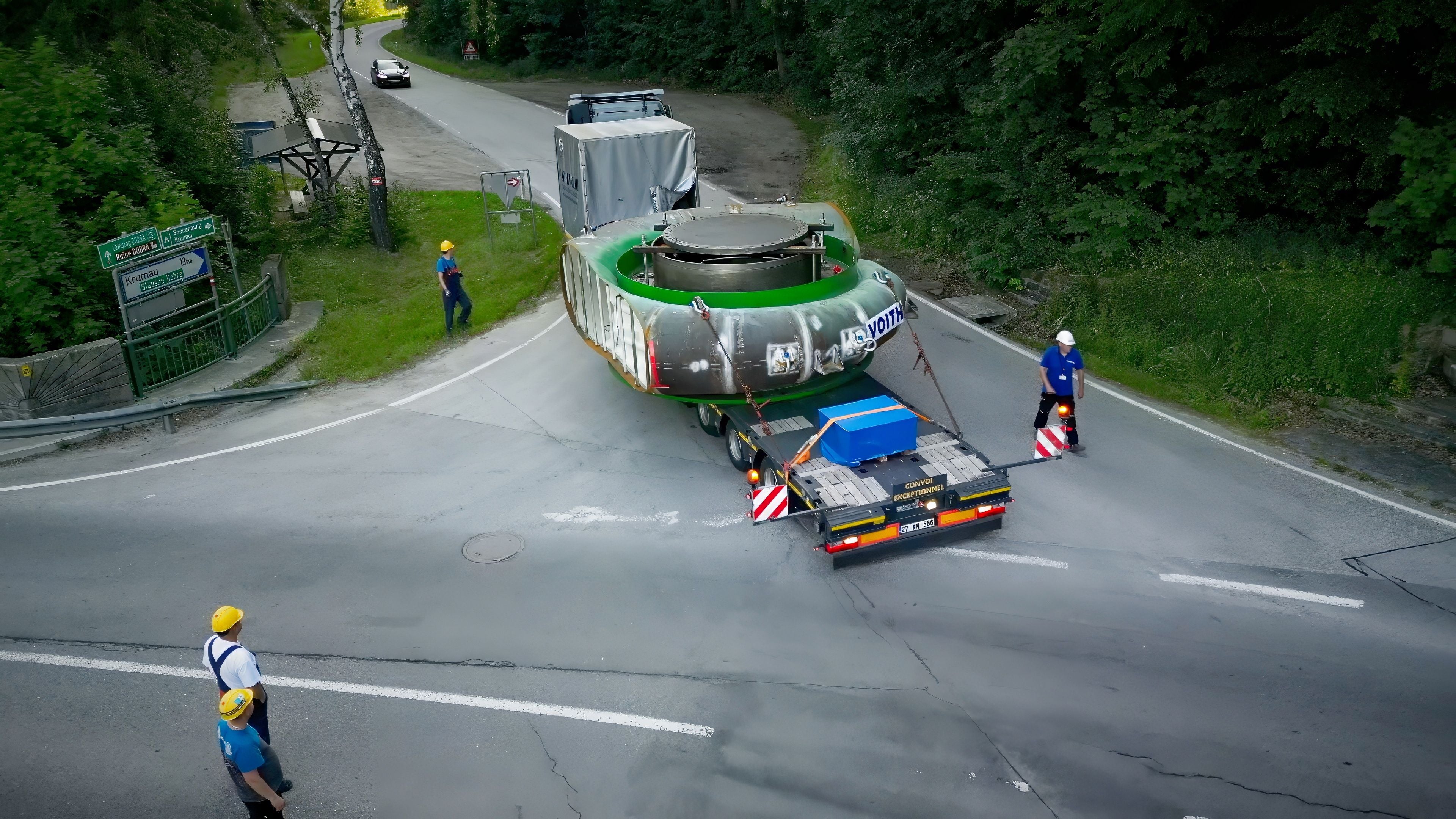 Schwertransport mit großer Pumpenspirale auf Tieflader, mehrere Arbeiter sichern die Straße.