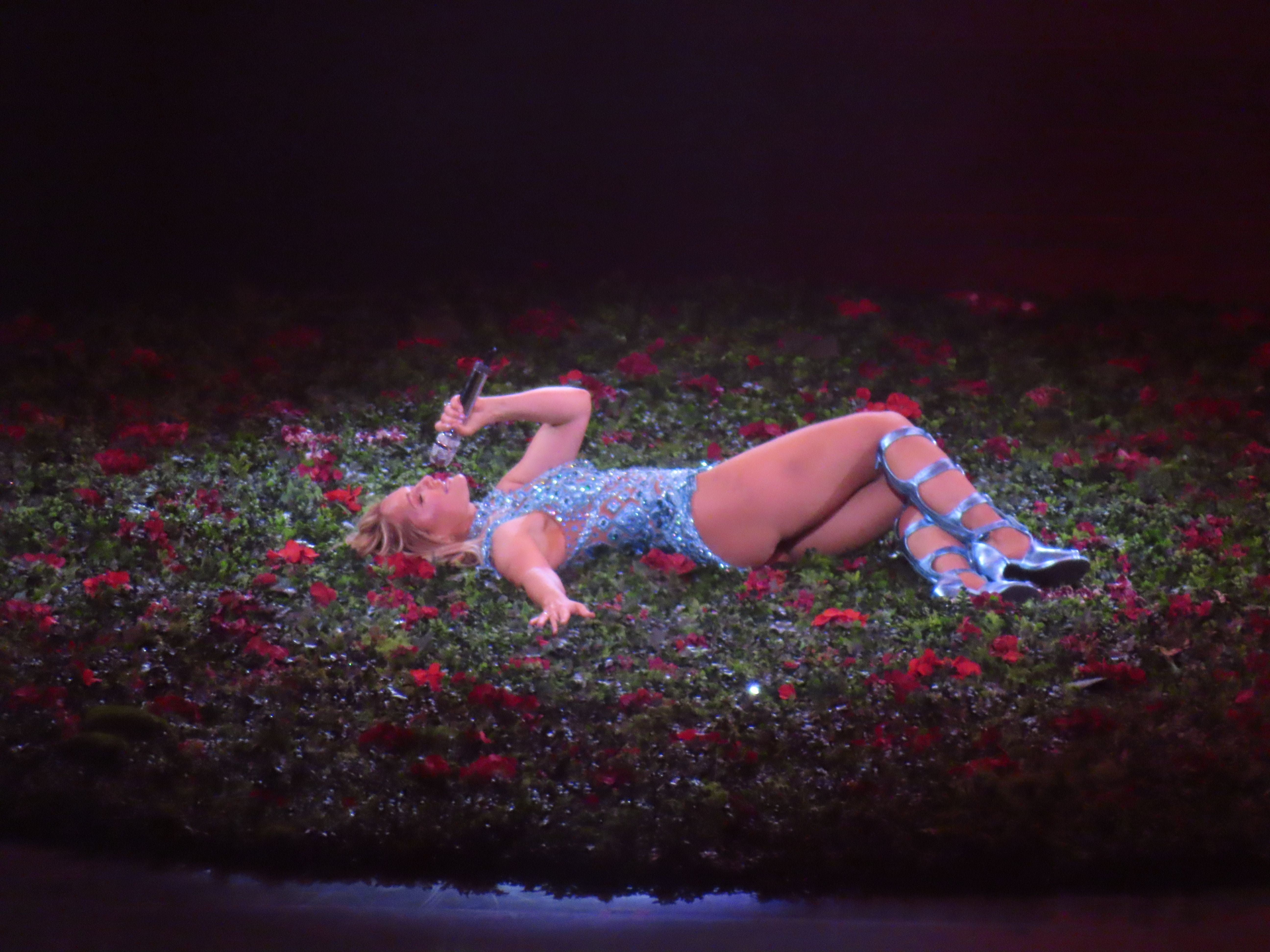 Frau im glitzernden Kleid liegt singend mit Mikrofon auf einer blumengeschmückten Bühne.