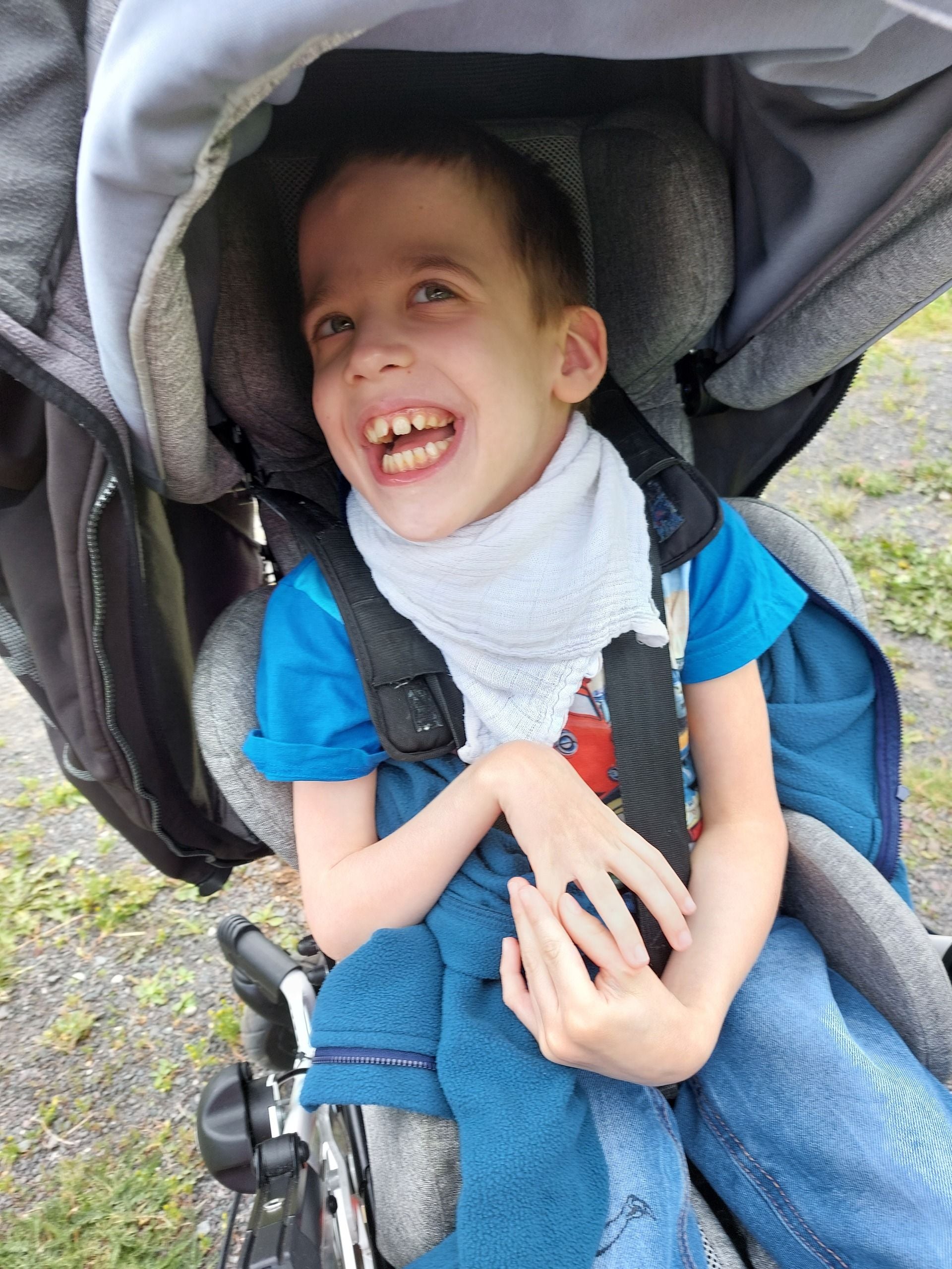 Ein lachender Junge sitzt in einem Kinderwagen und trägt ein blaues Shirt und eine Decke.