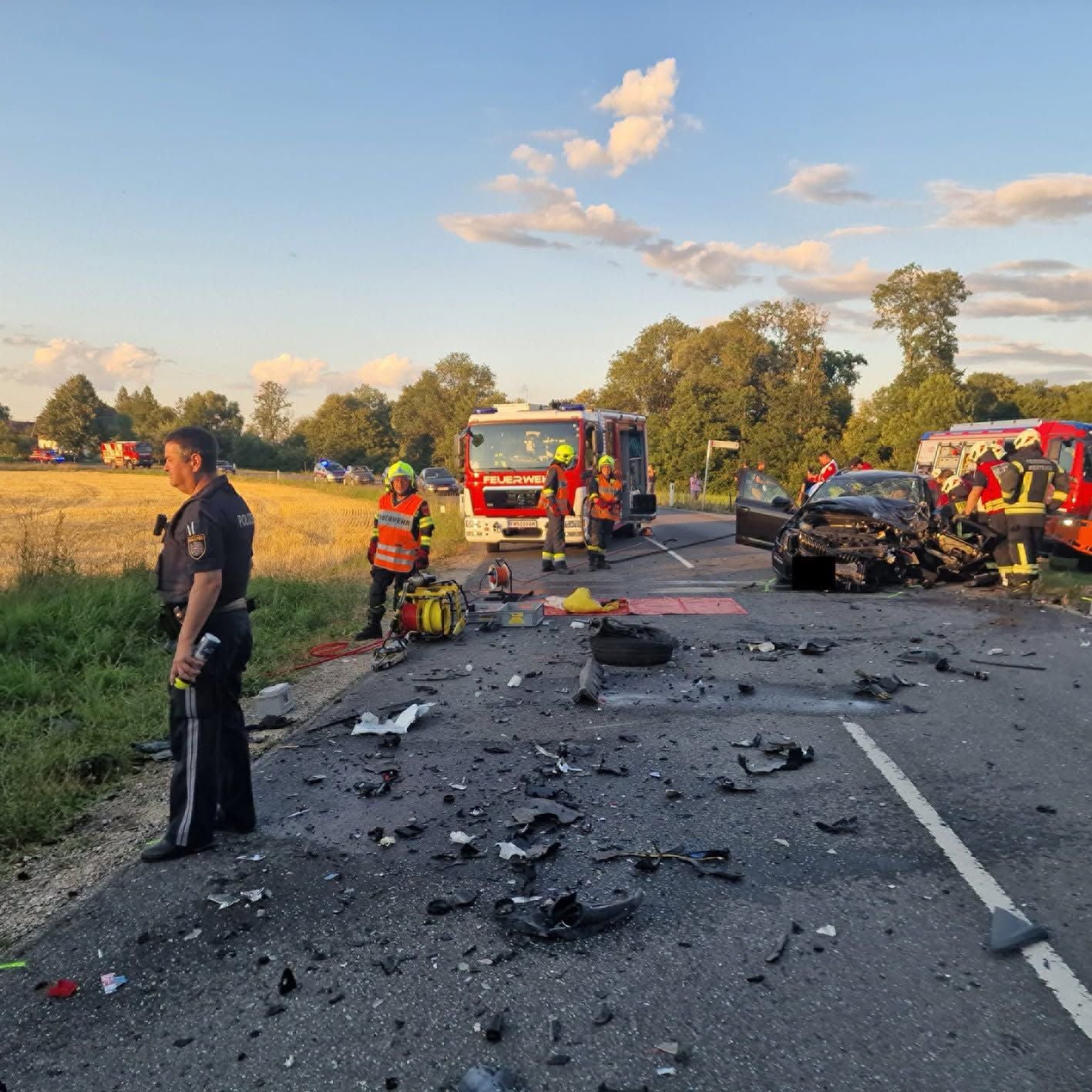 Verkehrsunfall mit Trümmern, Polizei und Feuerwehrleuten auf Landstraße bei Sonnenuntergang.