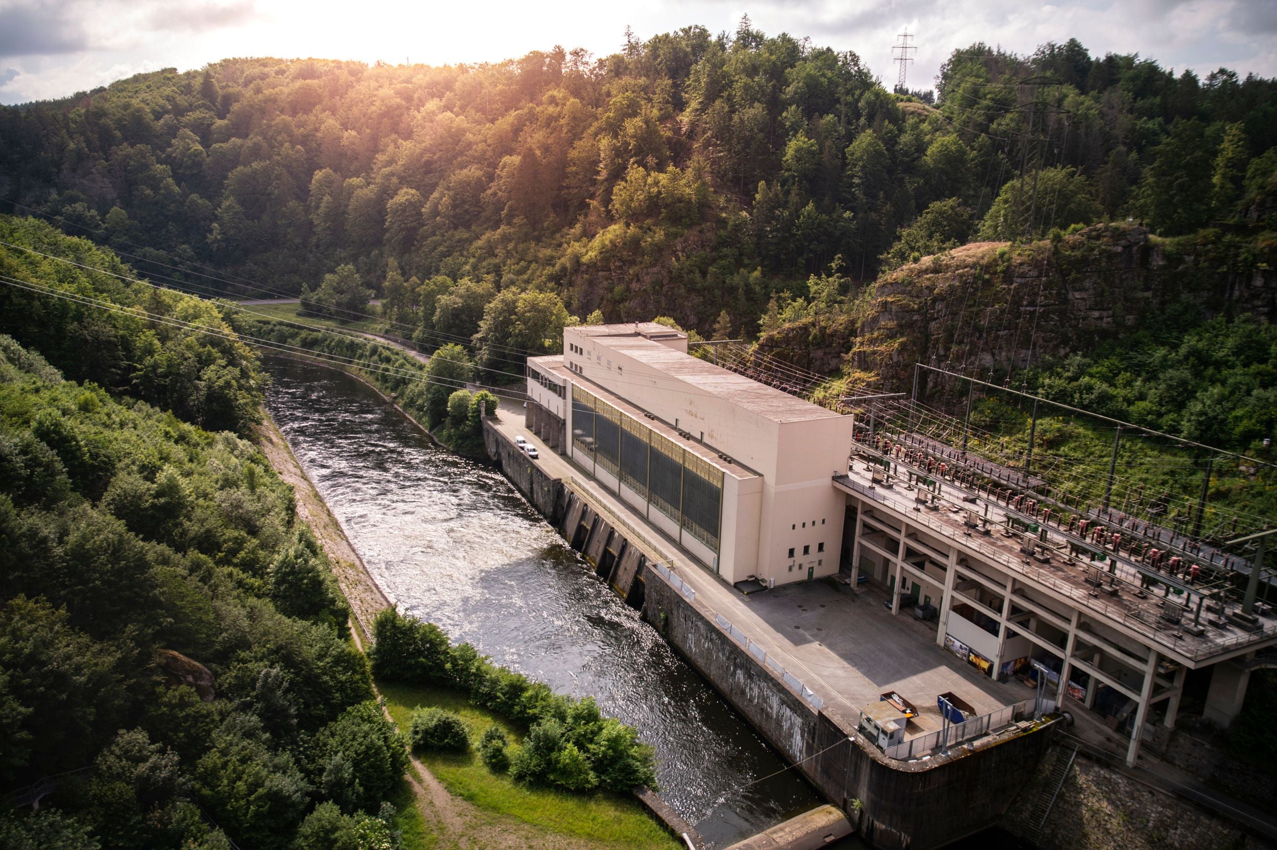 Luftaufnahme eines Wasserkraftwerks an einem Fluss, umgeben von bewaldeten Hügeln bei Sonnenlicht.
