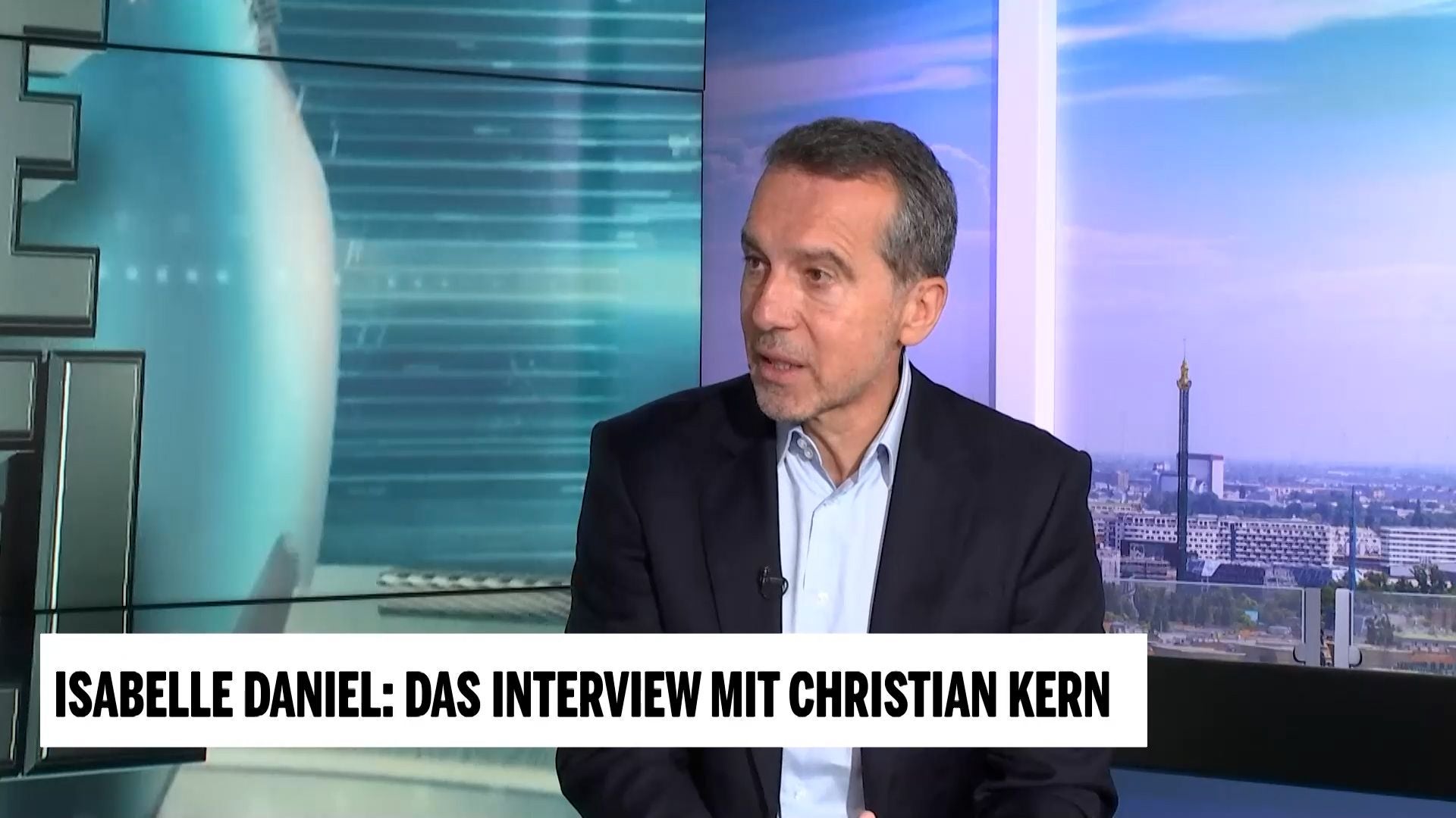 Ein Mann im Anzug sitzt im Fernsehstudio für ein Interview mit Stadtblick im Hintergrund.
