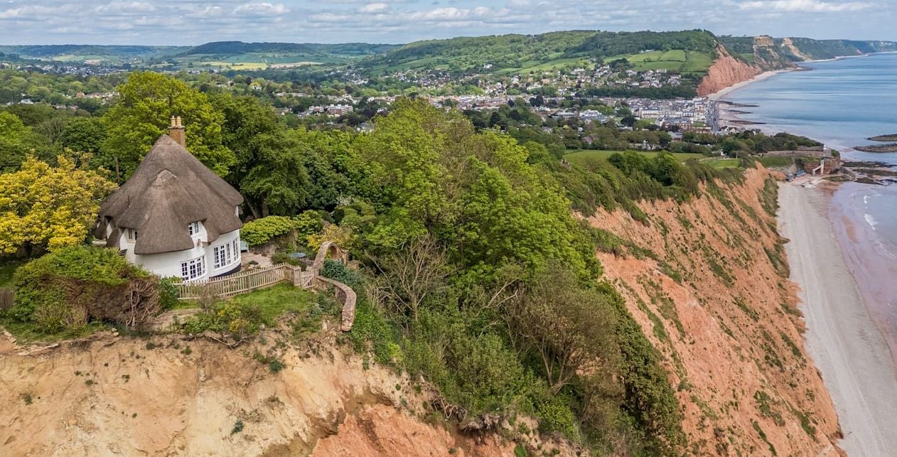 Reetgedecktes Haus auf einer Klippe mit Blick auf Meer, Strand und eine kleine Stadt im Hintergrund.