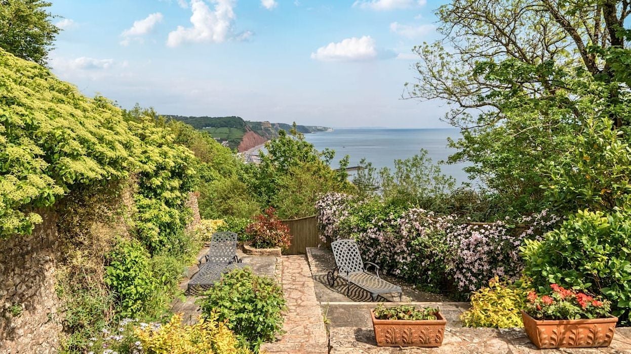 Terrasse mit Liegestühlen, umgeben von Blumen und Büschen, mit Meerblick im Hintergrund.