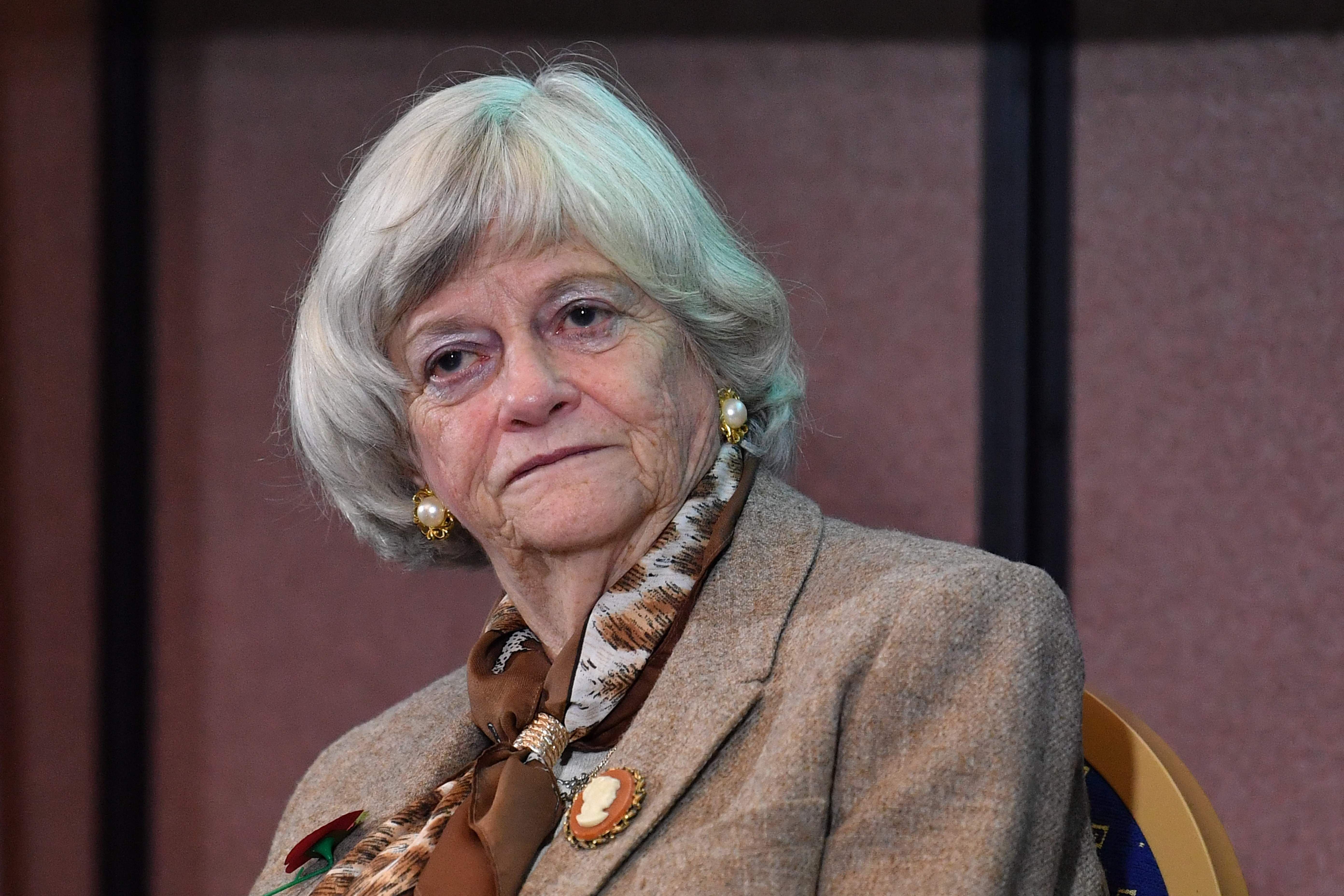 Ältere Frau mit grauen Haaren, beiger Jacke und Halstuch sitzt nachdenklich auf einem Stuhl.