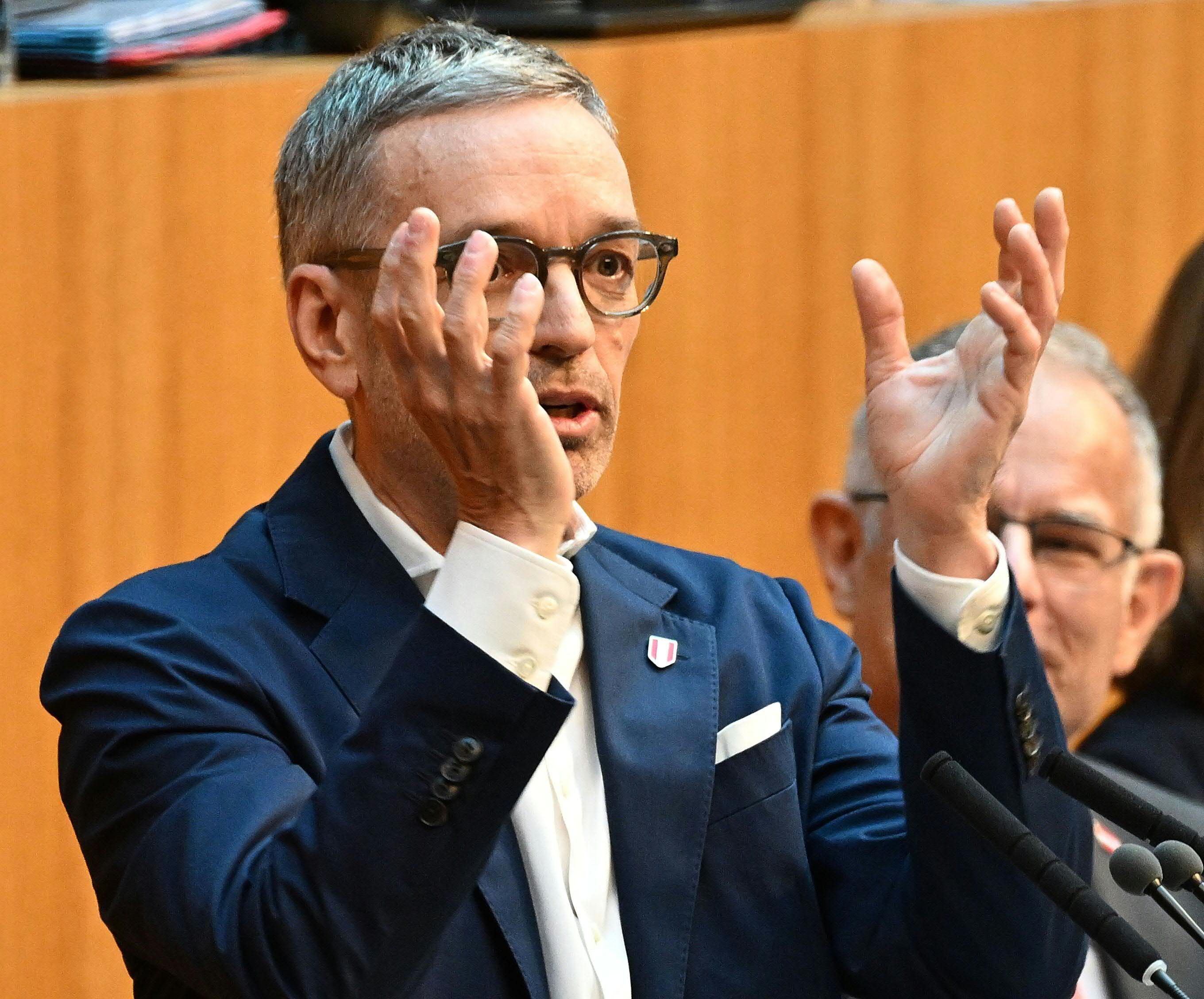 Ein Mann im Anzug spricht gestikulierend in einem Plenarsaal vor Mikrofonen.