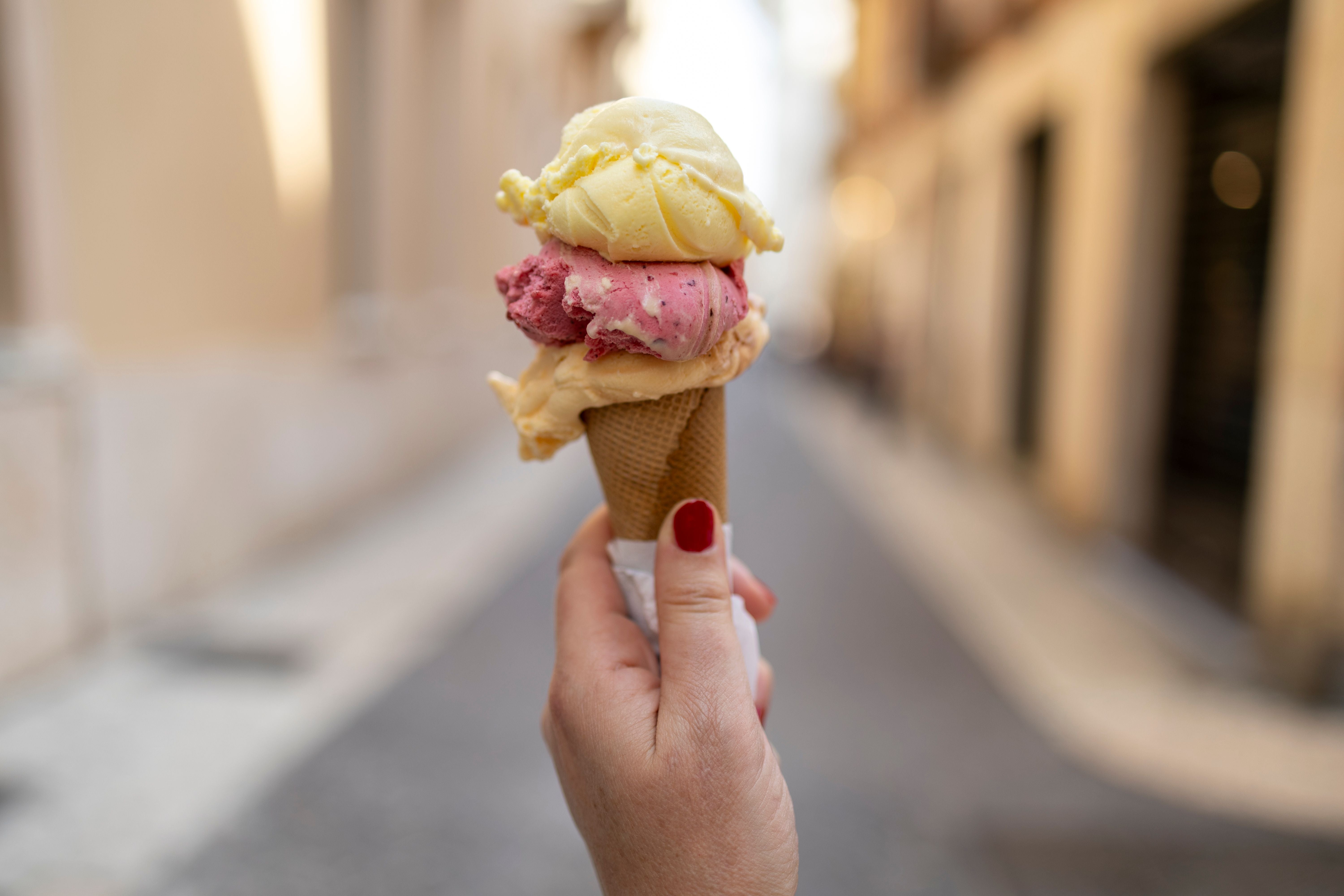 Hand hält Eiswaffel mit drei Kugeln Eis auf unscharfer Straße, Eis beginnt zu schmelzen.