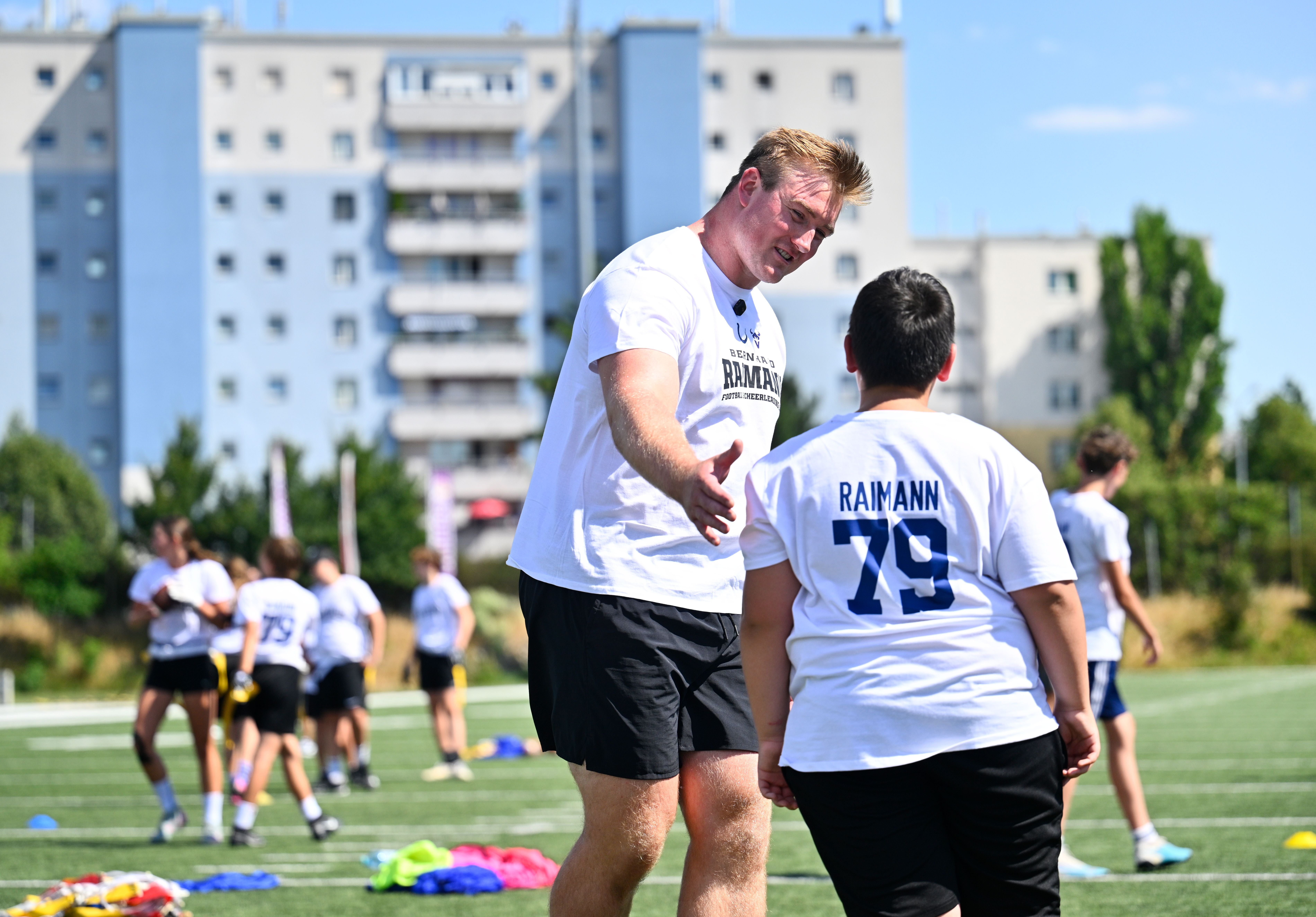 Ein Mann begrüßt einen Jungen auf einem Sportplatz bei einem Football-Camp, andere trainieren im Hintergrund.
