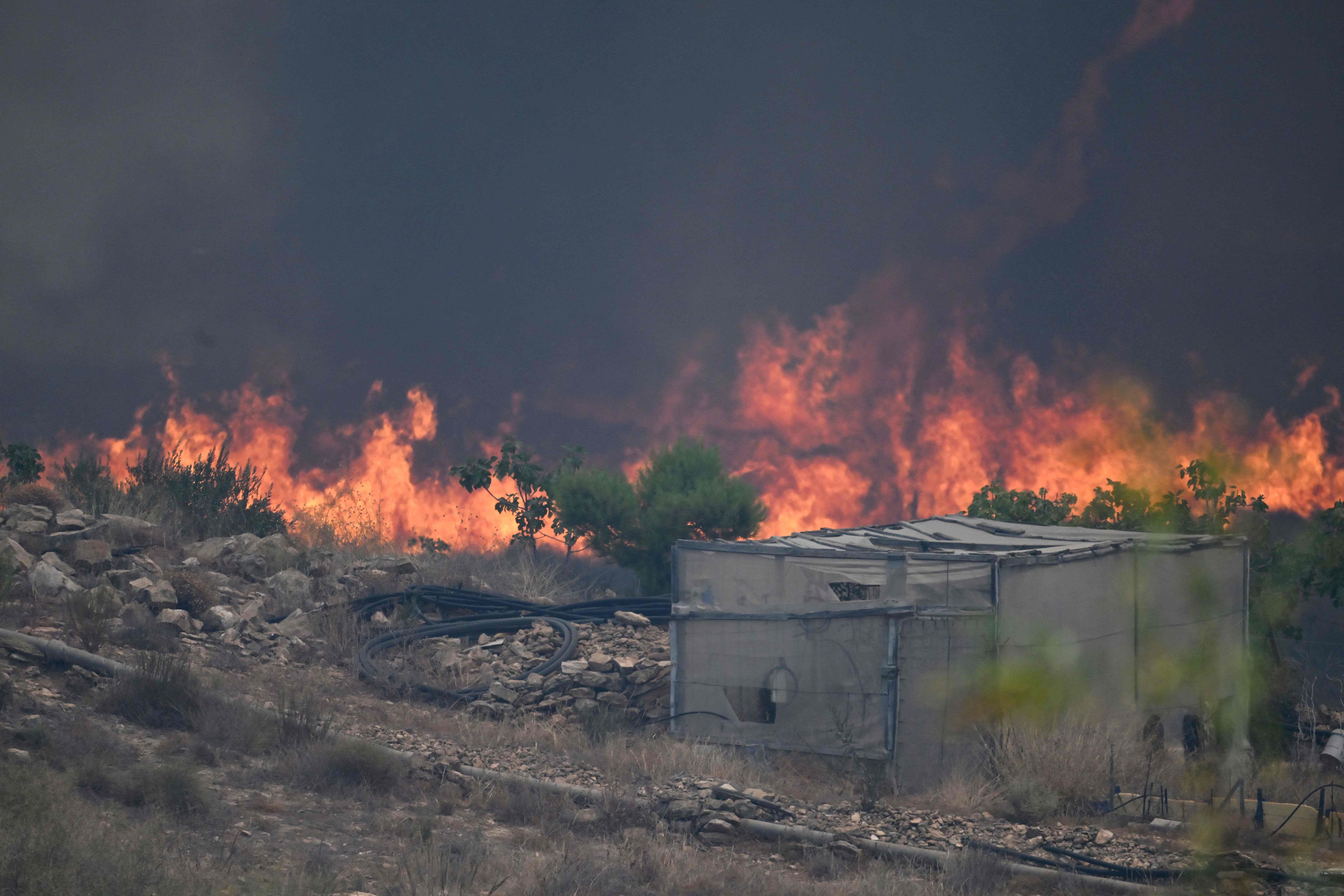 Ein großes Feuer wütet hinter einem Gebäude in einer trockenen, steinigen Landschaft.