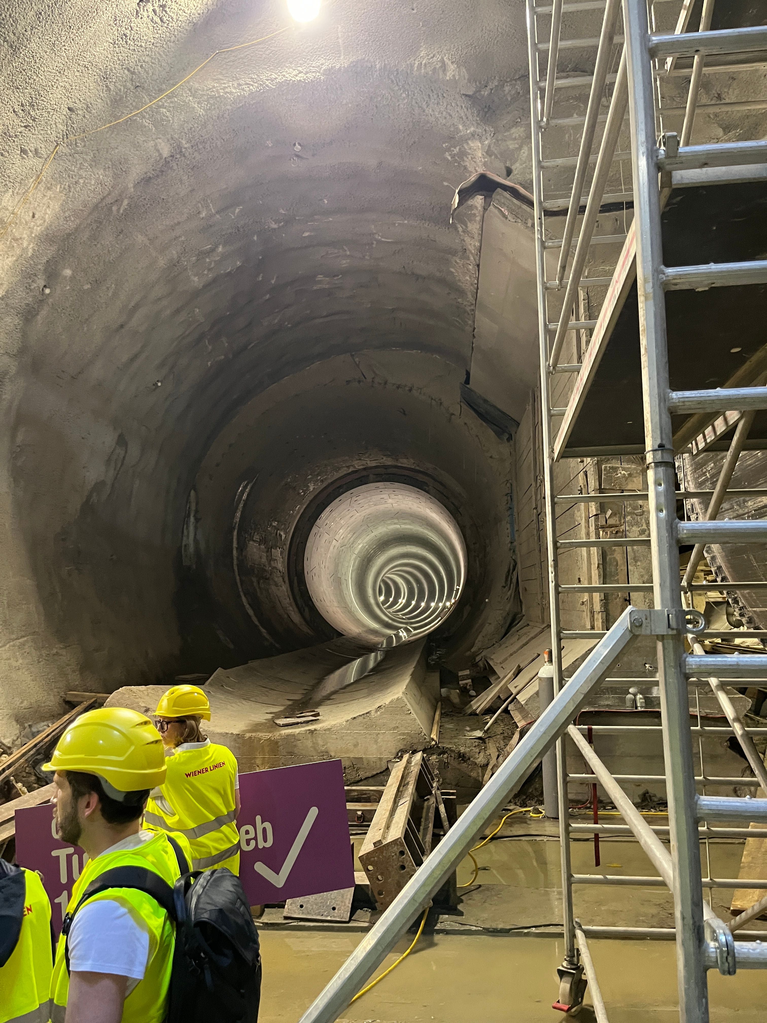 Personen mit Schutzhelmen in einem Tunnelbau, rechts Baugerüst, im Hintergrund Tunnelröhre.