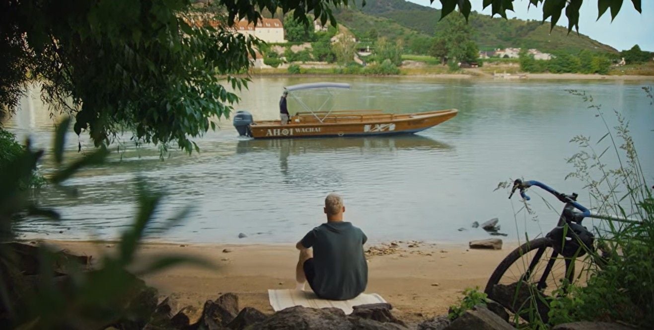 Arnautović sitzt im Video am Donauufer, im Hintergrund fährt ein Holzboot vorbei, rechts steht ein Fahrrad.