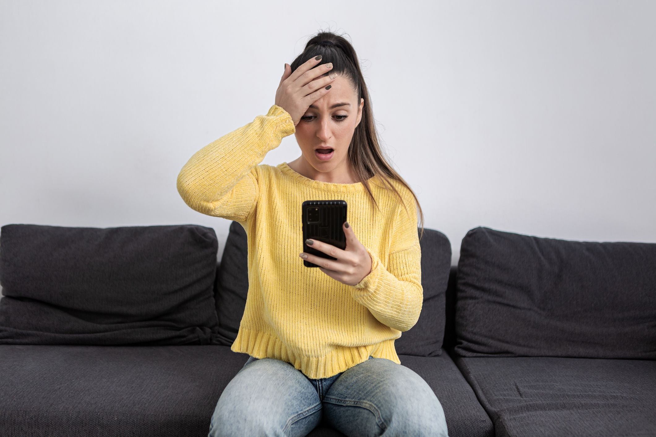 Junge Frau im gelben Pullover schaut erschrocken auf ihr Handy und fasst sich an die Stirn.