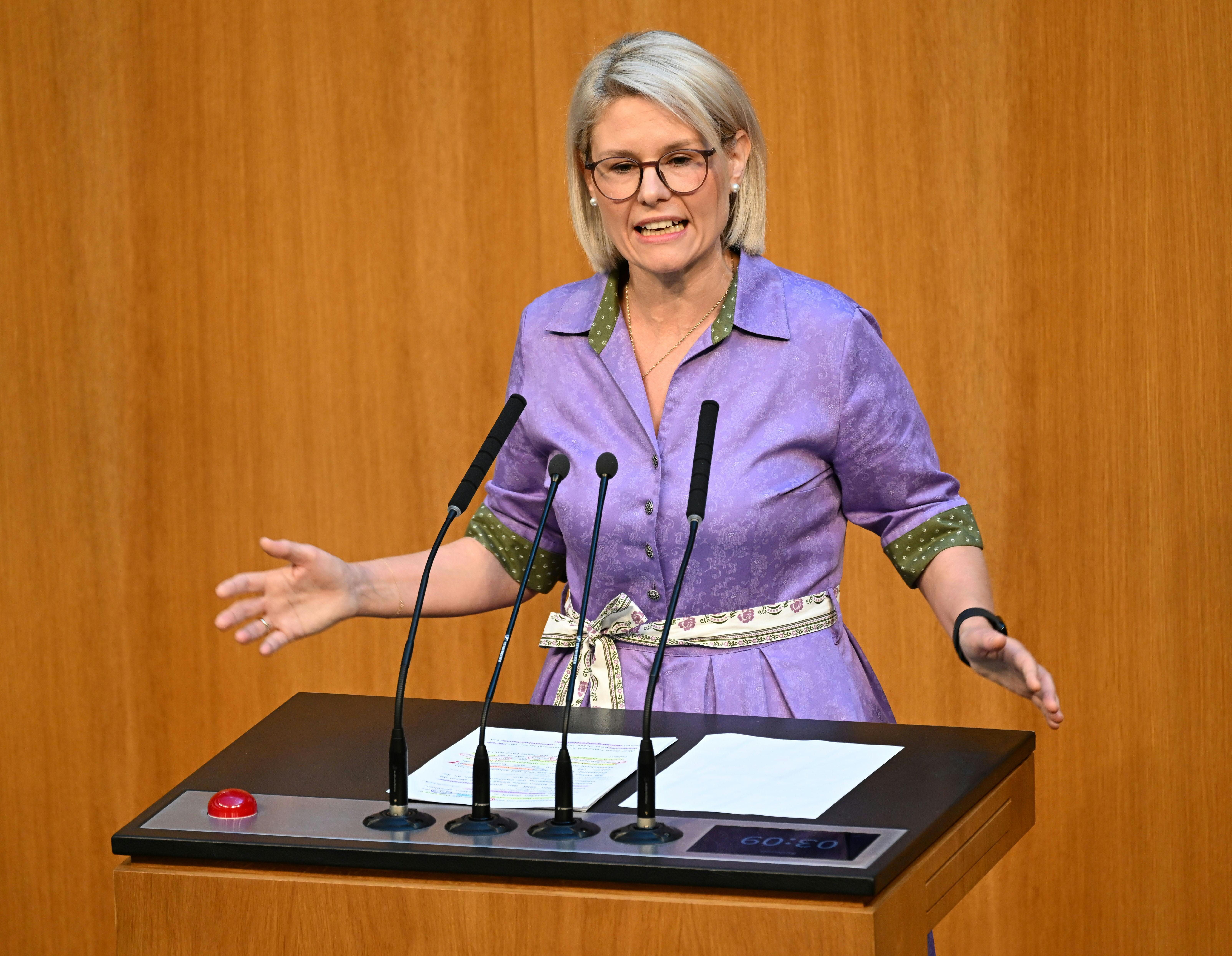 Frau spricht am Rednerpult im Parlament, trägt lila Kleid und Brille, gestikuliert mit den Händen.