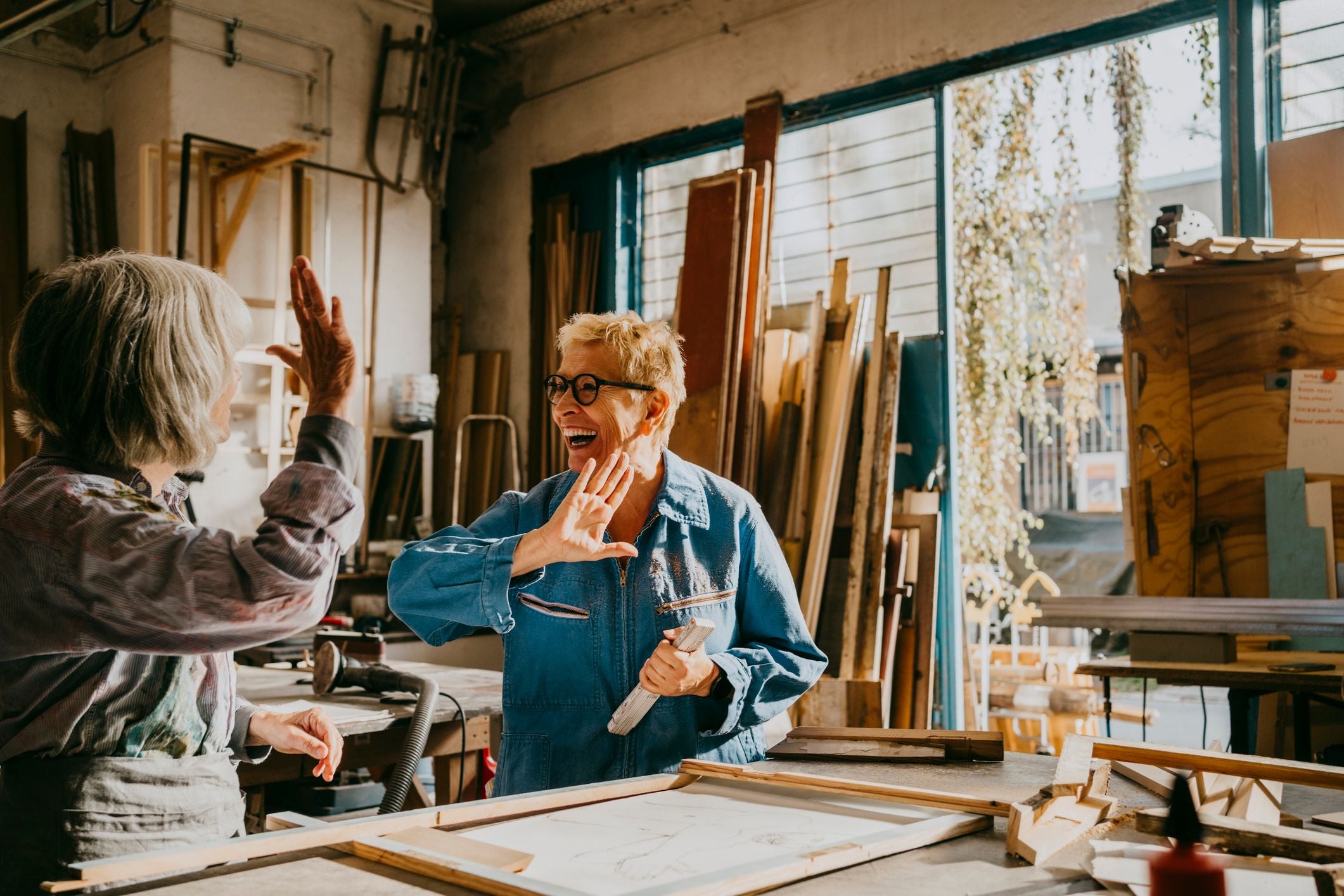 Zwei ältere Frauen geben sich ein High-Five in einer sonnigen Holzwerkstatt.