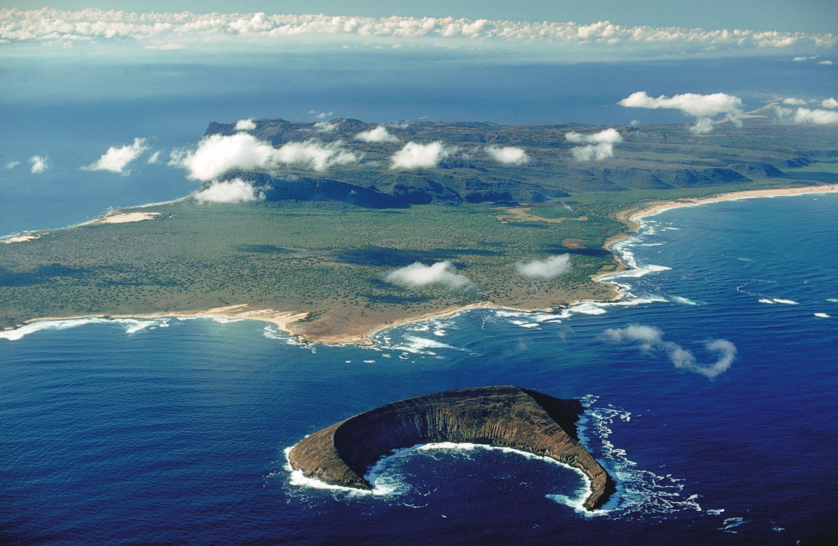 Luftaufnahme von Niihau, einer grünen Insel mit Küste und vorgelagertem Krater im blauen Meer.