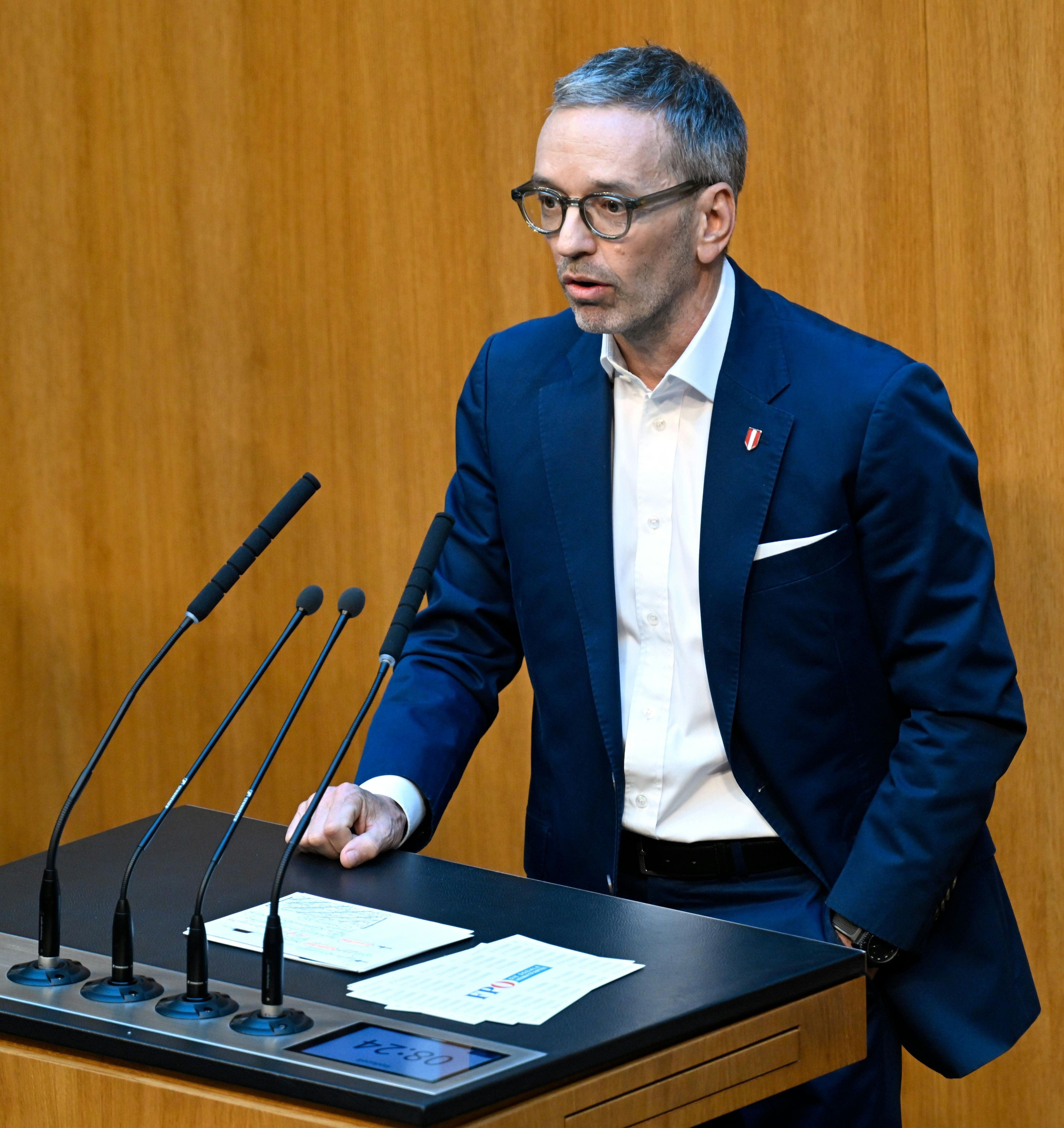Ein Mann im Anzug spricht an einem Rednerpult mit mehreren Mikrofonen im Parlament.