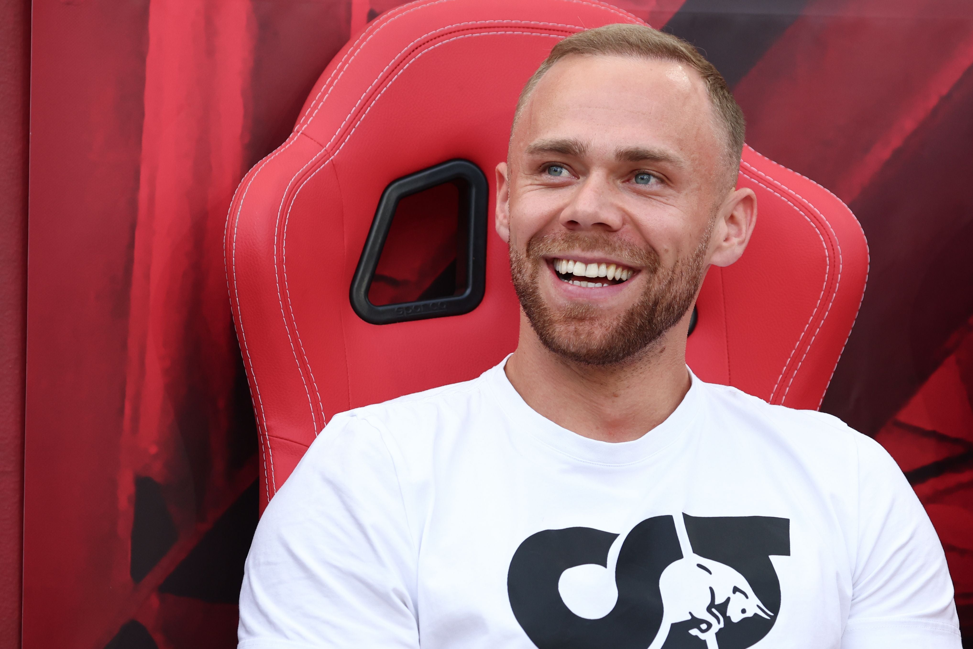 Ein lächelnder Mann sitzt auf einem roten Sportsitz vor rotem Hintergrund.