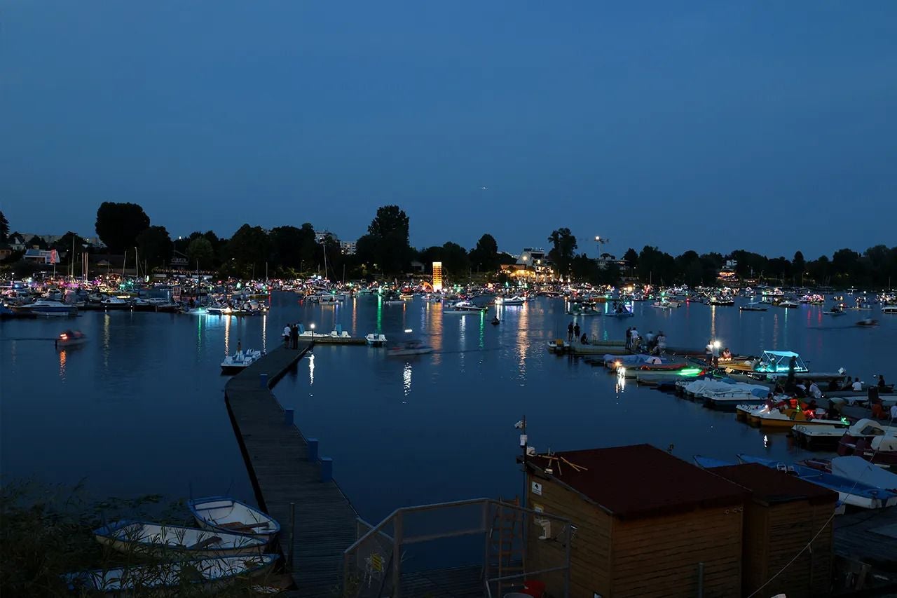 Viele beleuchtete Boote liegen abends an Stegen der Alten Donau, Menschen feiern am Ufer.