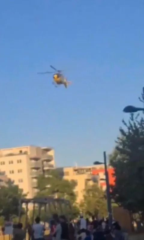 Ein gelber Rettungshubschrauber fliegt über einen Park mit Menschen und Wohnhäusern im Hintergrund.