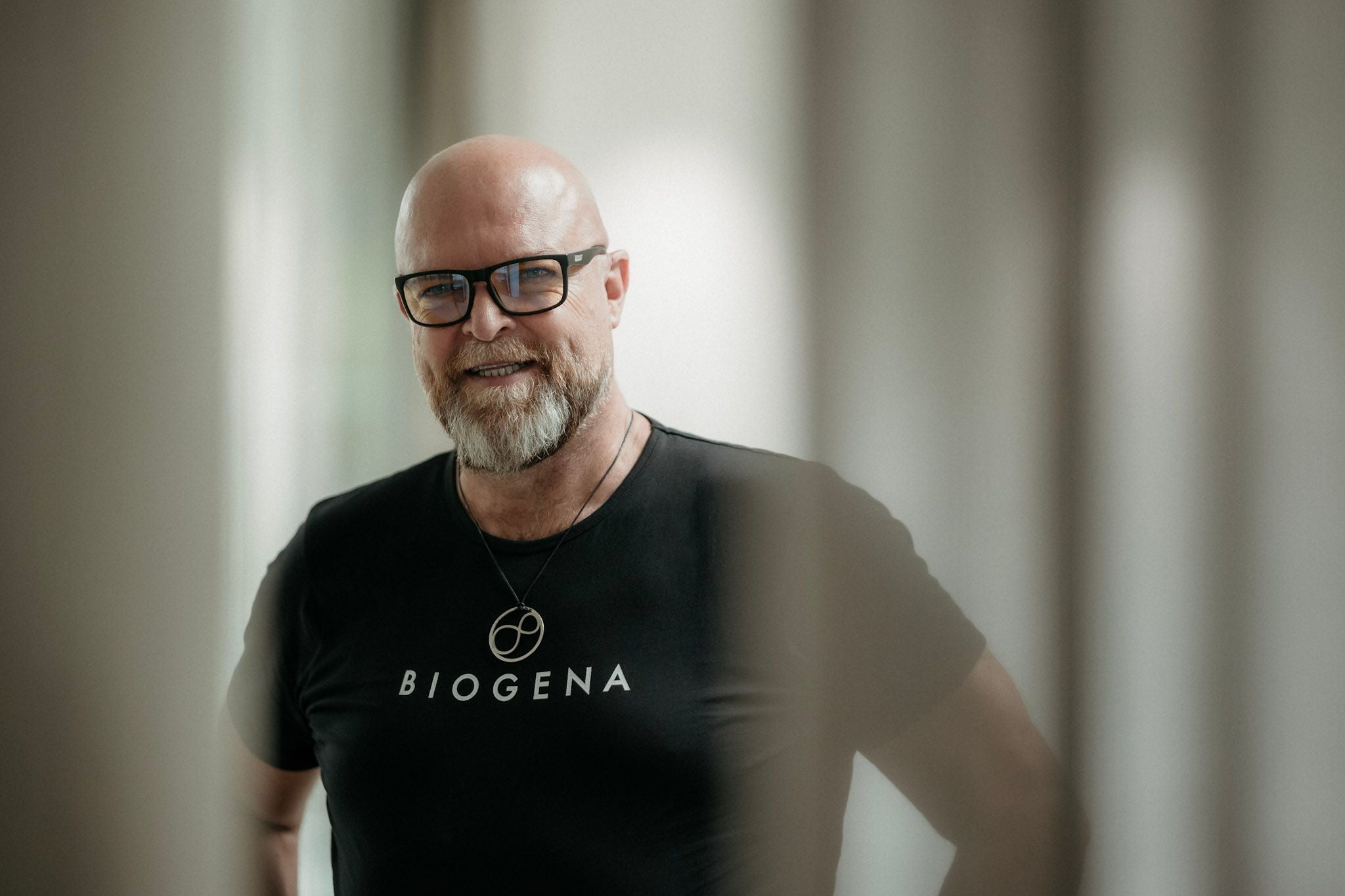 Ein lächelnder, glatzköpfiger Mann mit Brille und Bart trägt ein schwarzes „BIOGENA“-Shirt.