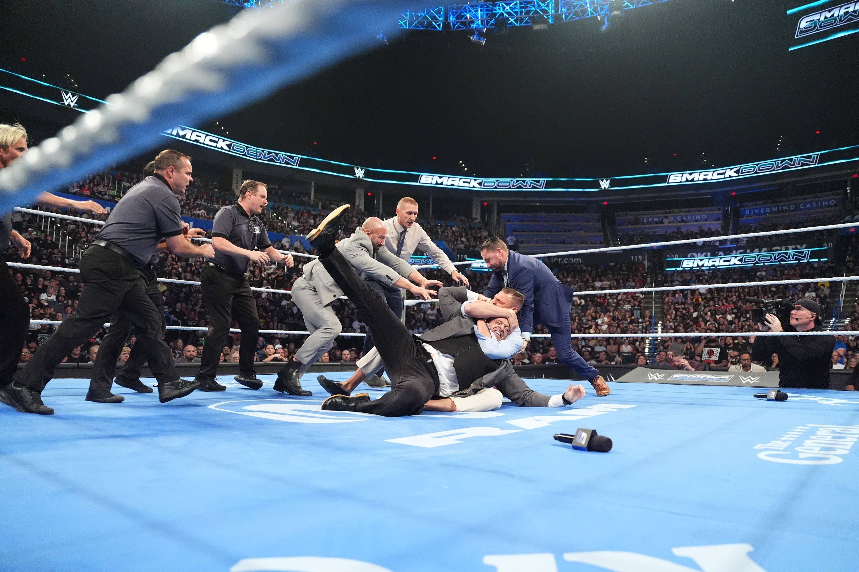 Mehrere Männer in Anzügen und Sicherheitsleute kämpfen im Wrestling-Ring vor großem Publikum.