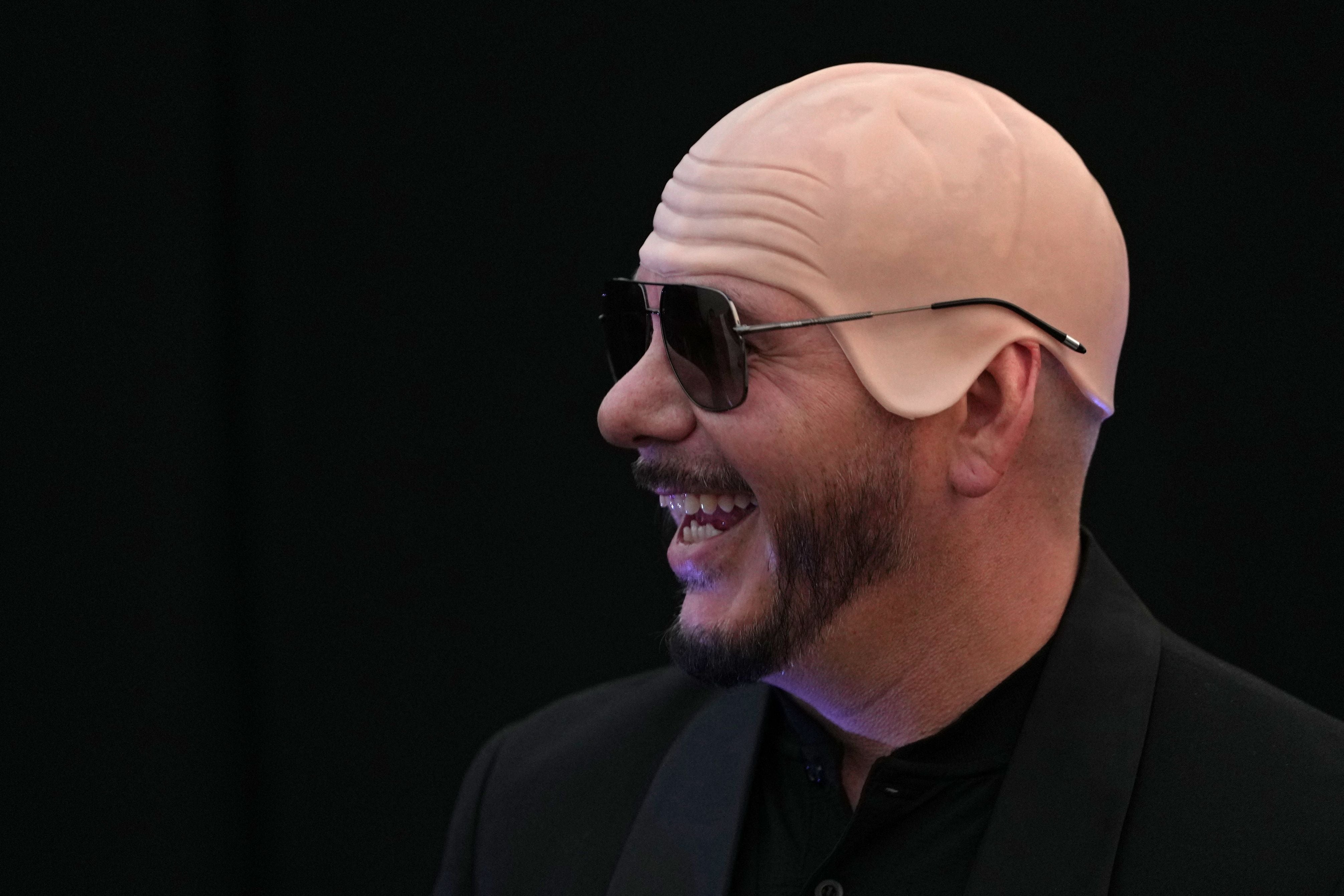 Ein lachender Mann mit Sonnenbrille trägt eine Glatze-Perücke vor schwarzem Hintergrund.