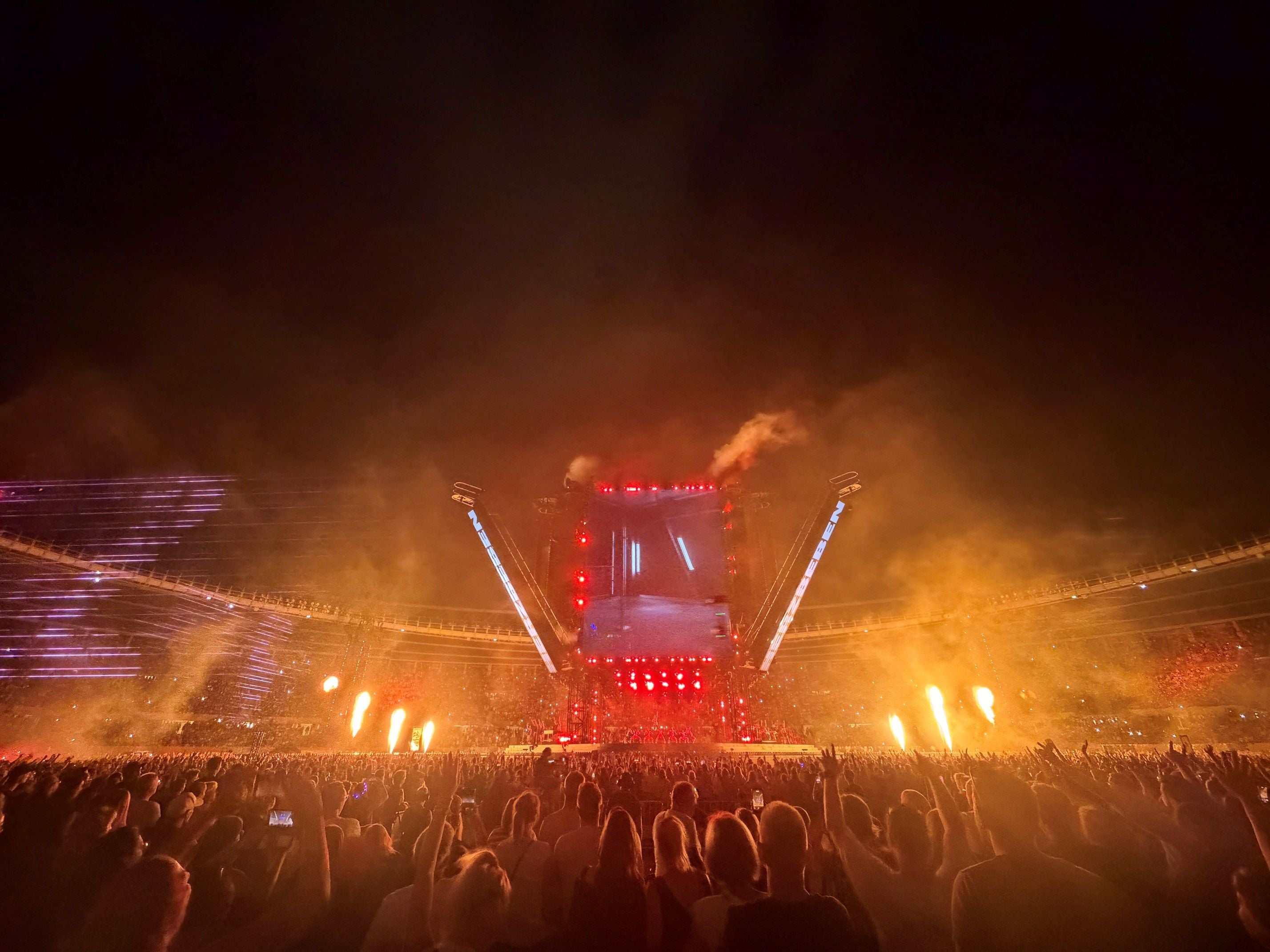 Großes Open-Air-Konzert bei Nacht mit vielen Zuschauern, Feuer- und Lichteffekten auf der Bühne.