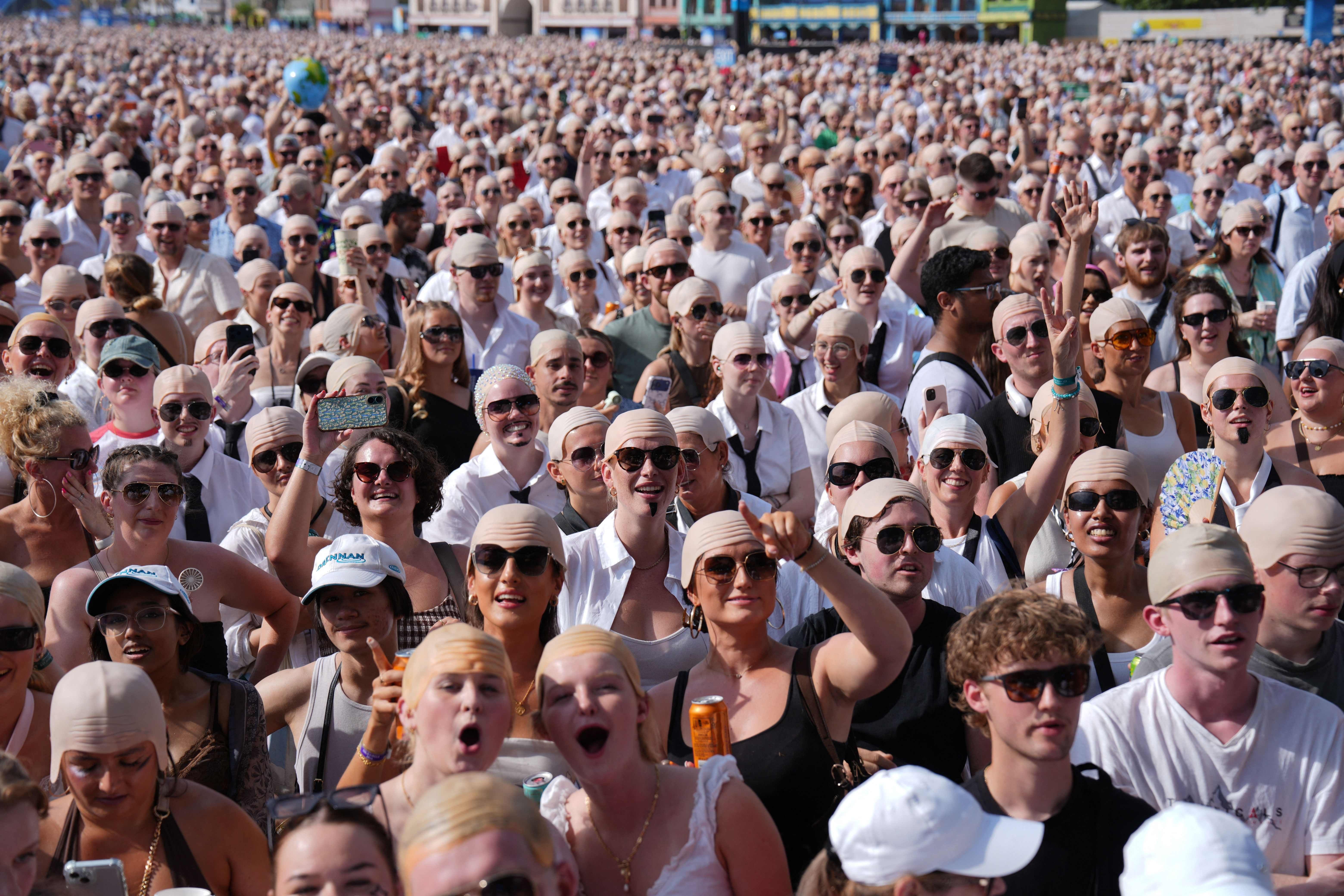 Eine große Menschenmenge trägt Glatzenhauben bei sonnigem Wetter vor einem Konzert im Freien.