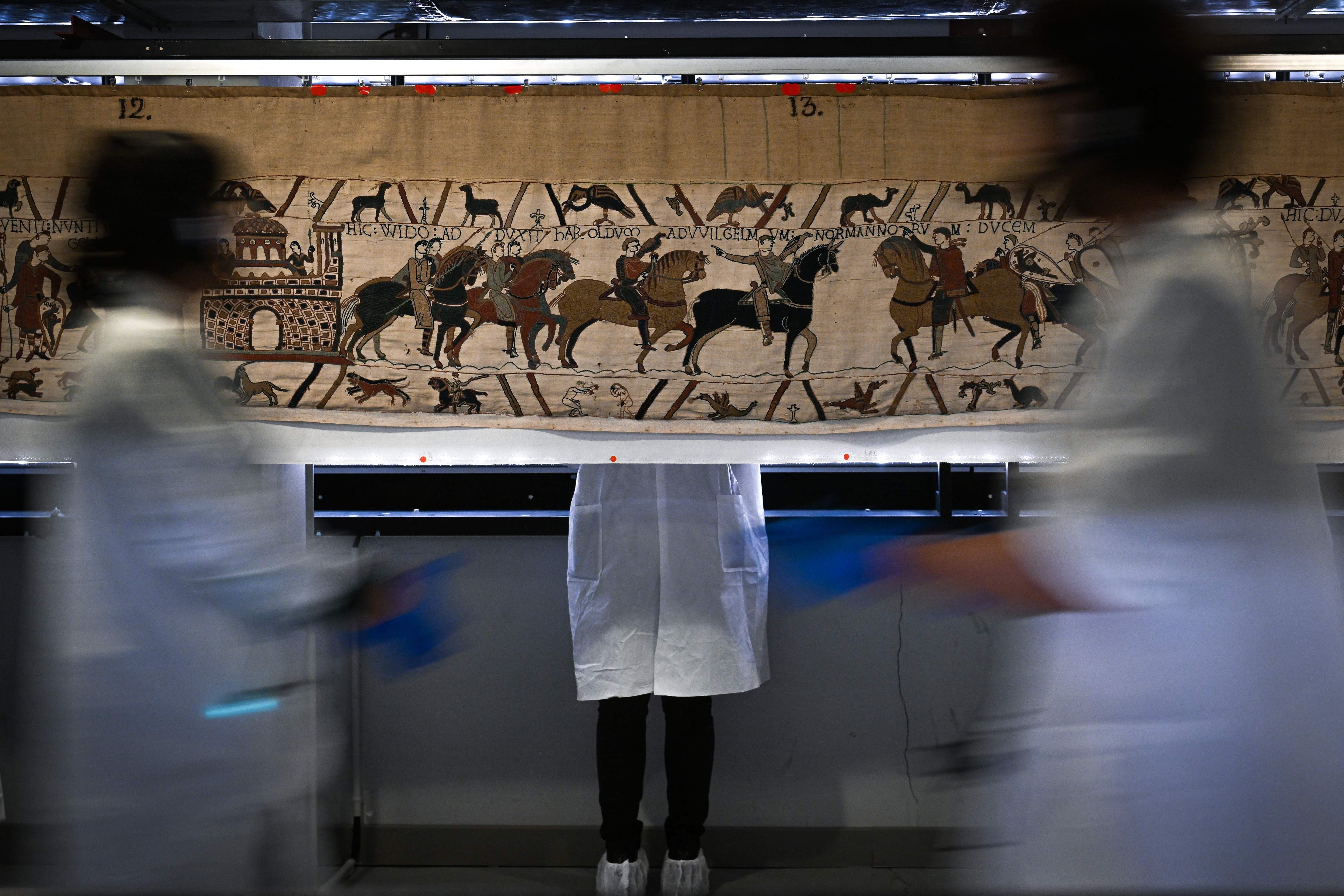 Menschen in weißen Kitteln stehen und gehen vor dem historischen Wandteppich von Bayeux.