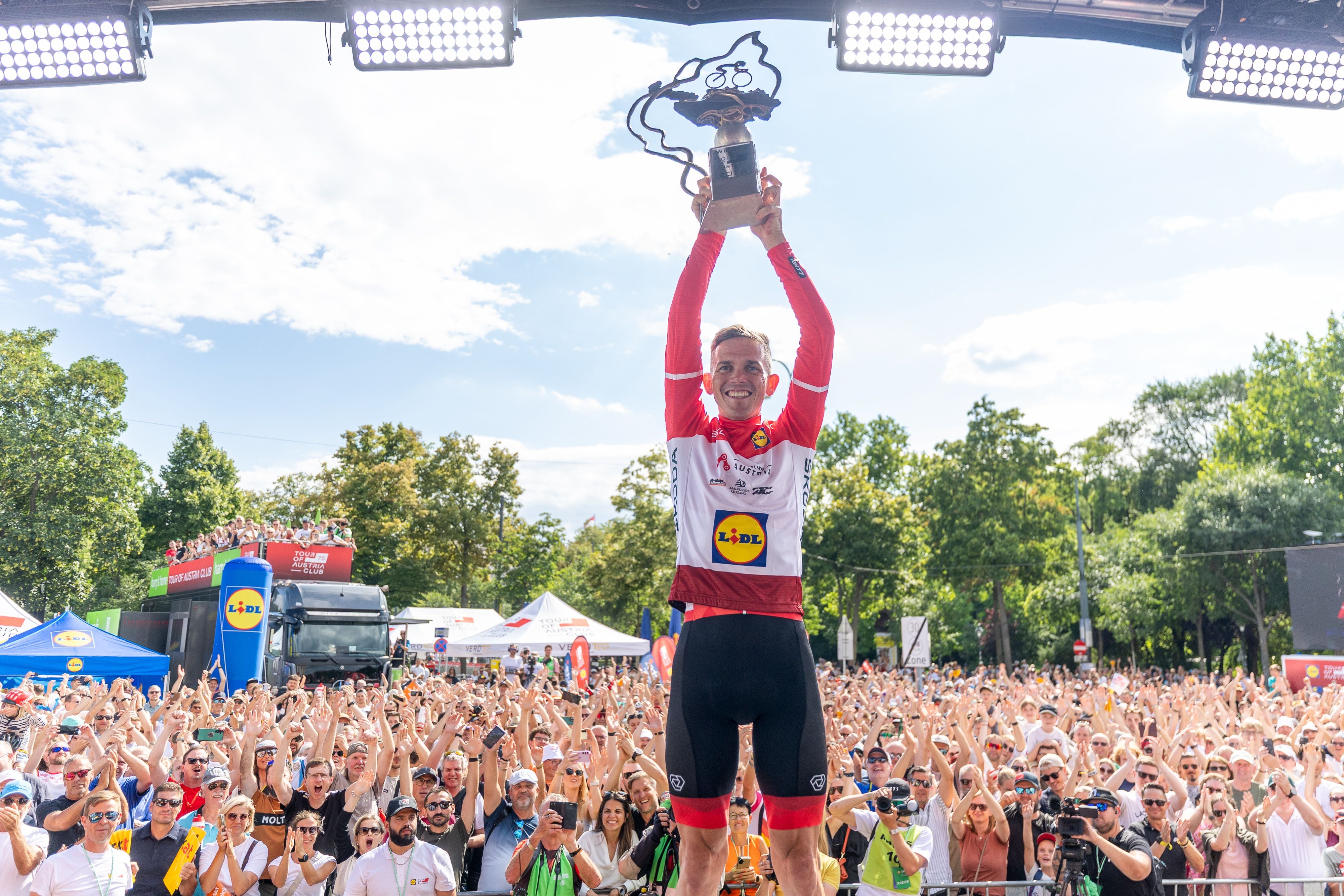 Radfahrer hebt Siegerpokal jubelnd vor einer großen Menschenmenge in Wien bei der Tour of Austria 2026.