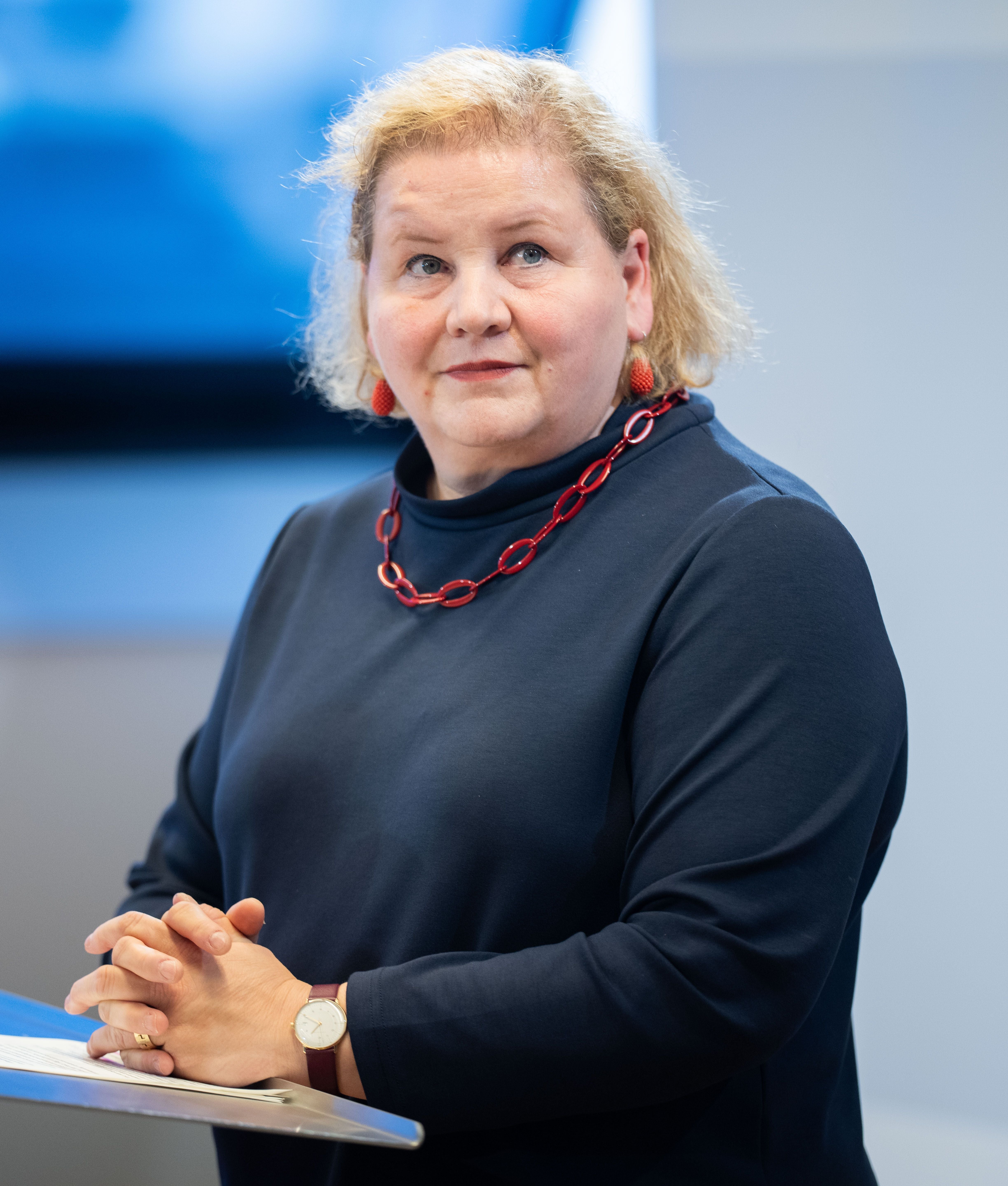 Frau mit blonden Haaren hält ein Statement, trägt dunkelblaues Oberteil und rote Kette.