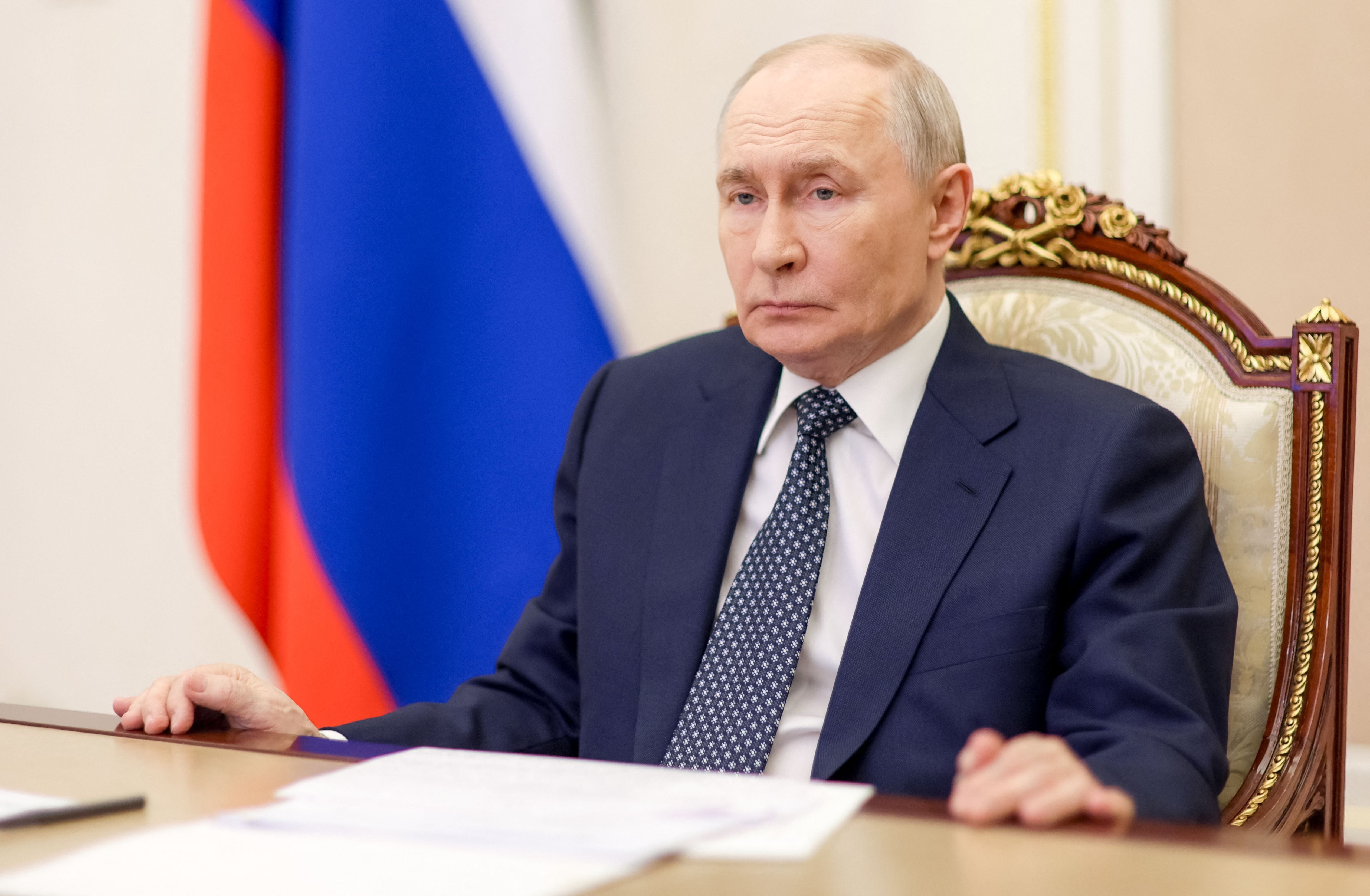 Ein Mann im Anzug sitzt an einem Tisch vor einer russischen Flagge und blickt ernst.