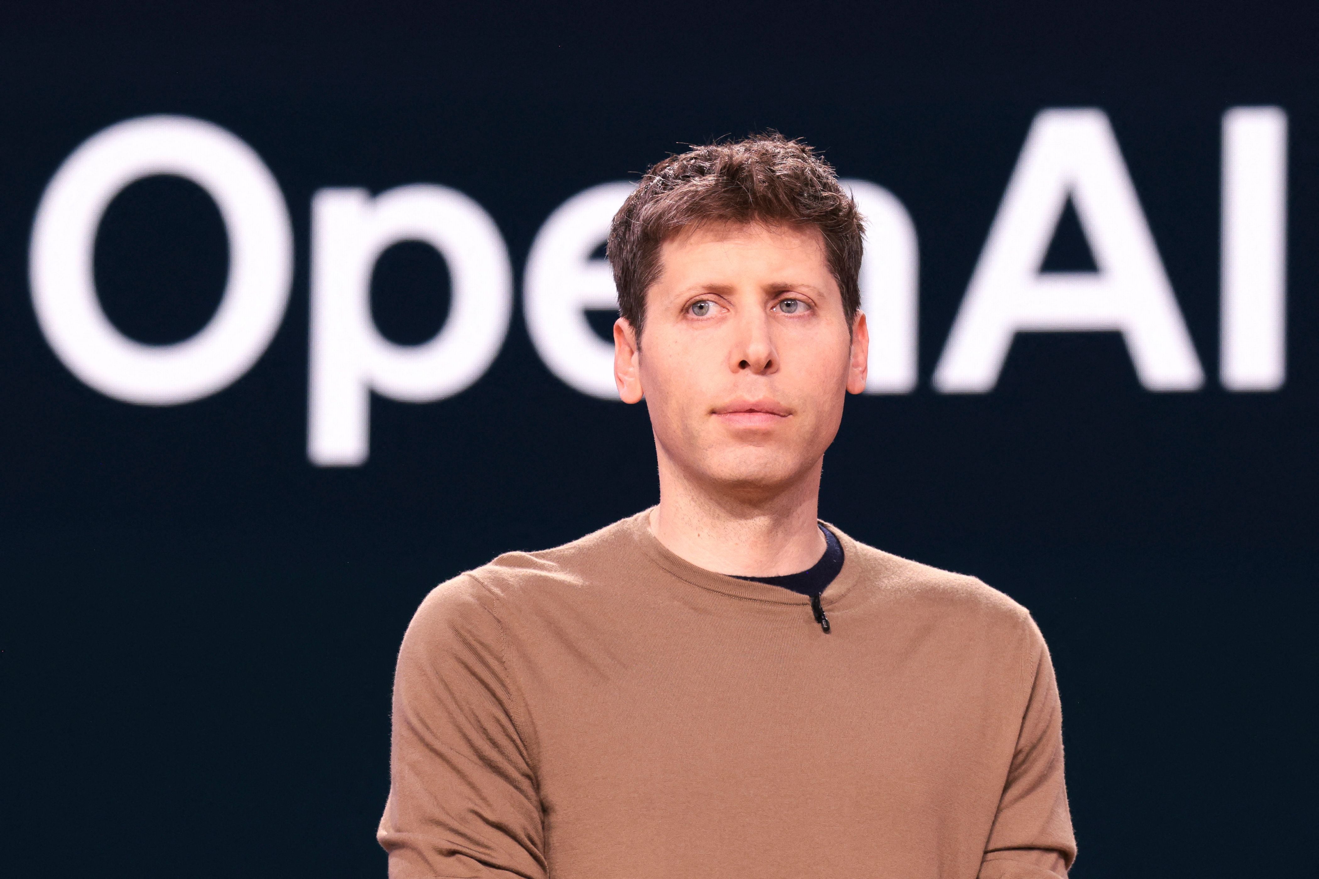 Ein Mann mit braunem Pullover steht vor einem Hintergrund mit dem Schriftzug „OpenAI“.