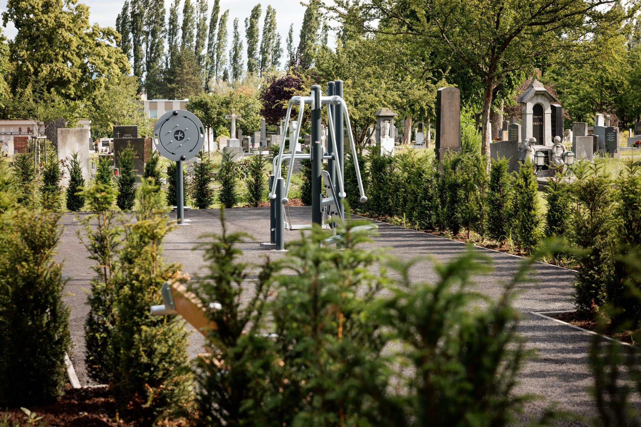 Outdoor-Fitnessgeräte stehen auf einem Weg zwischen Gräbern und Bäumen auf einem Friedhof.