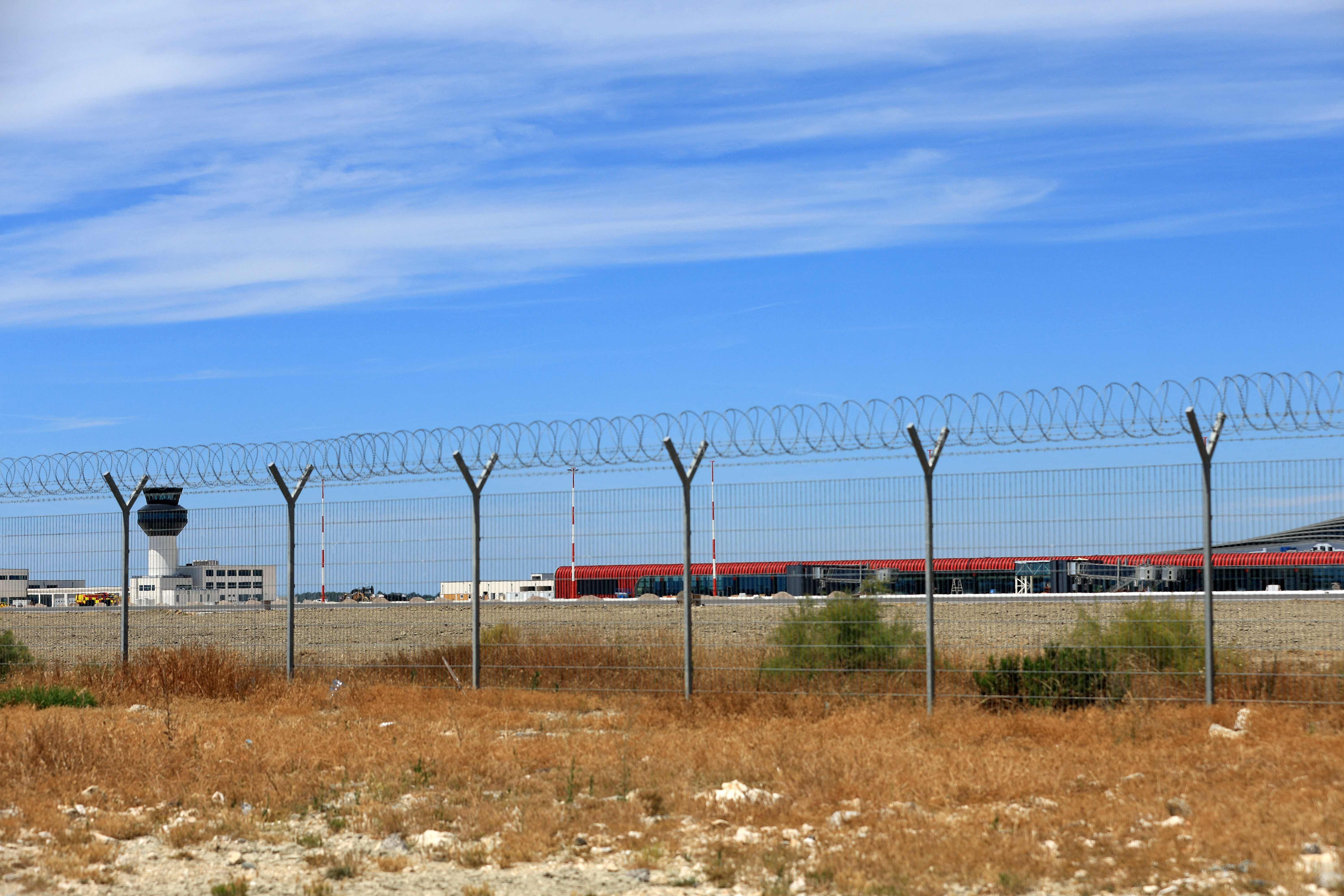 Weitblick auf einen neuen Flughafen hinter einem mit Stacheldraht gesicherten Zaun unter blauem Himmel.