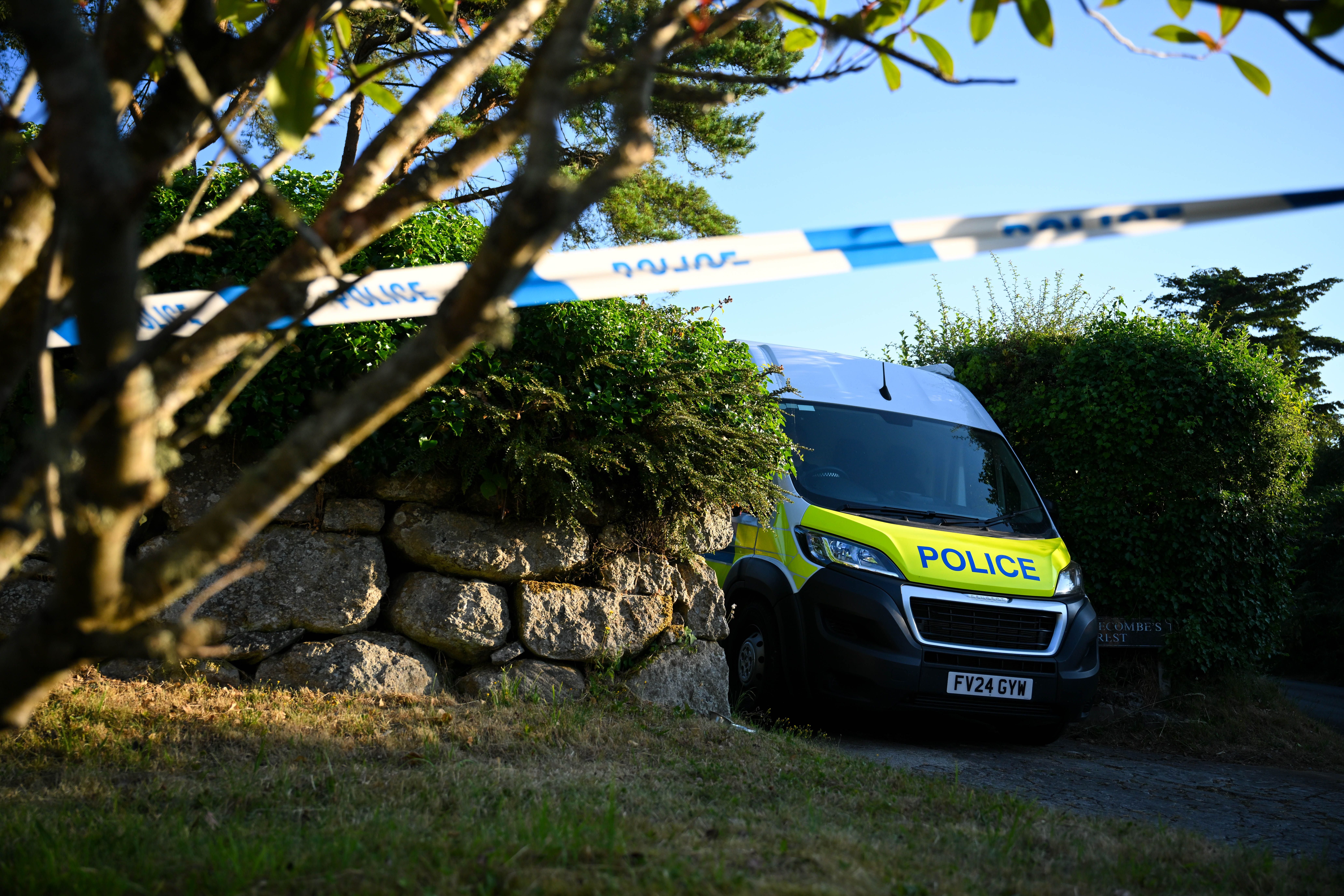 Ein Polizeiwagen steht hinter Absperrband neben einer Steinmauer und Büschen im Freien.