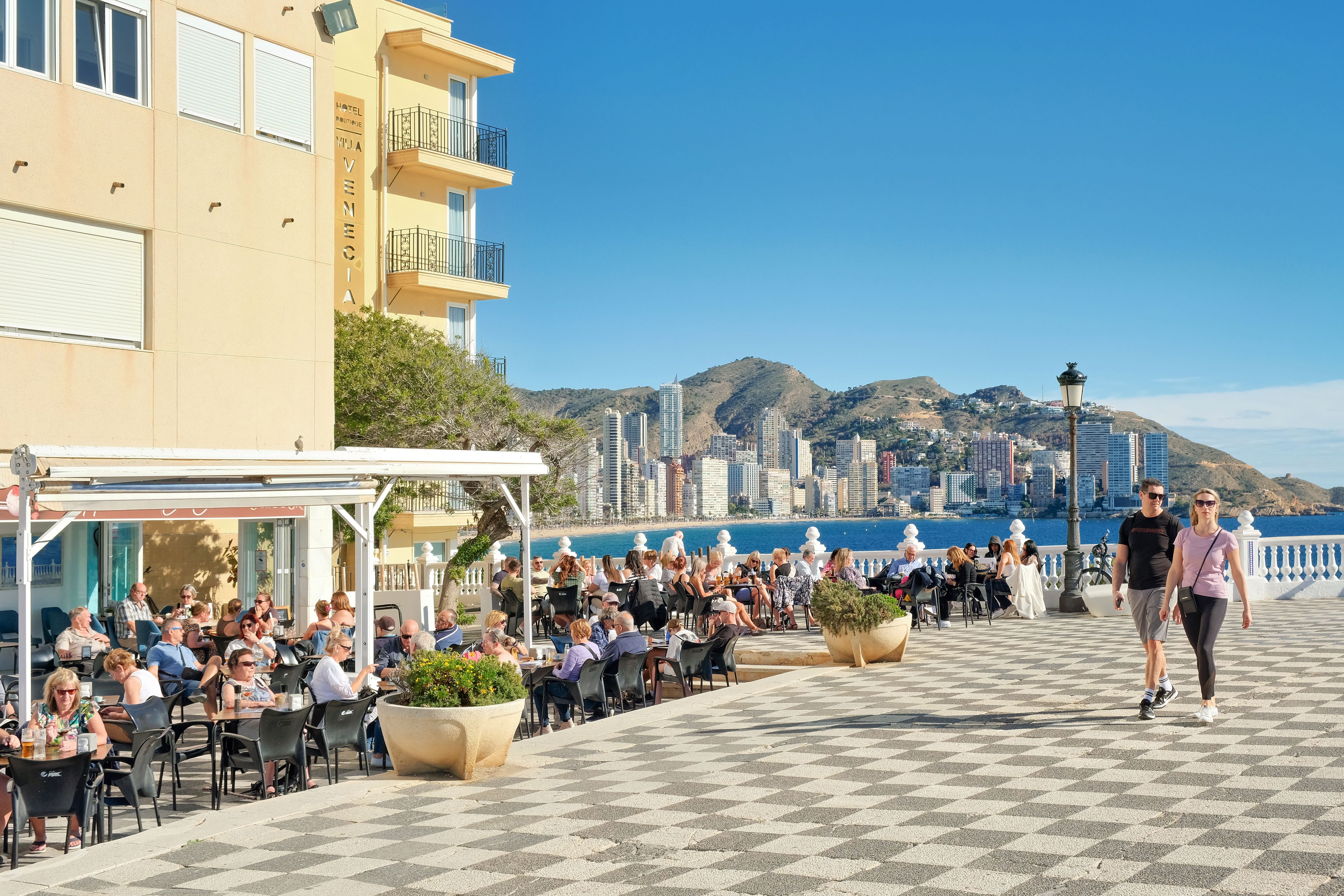 Menschen sitzen in Straßencafés und spazieren auf der Plaza de Santa Ana mit Blick auf Benidorms Skyline.