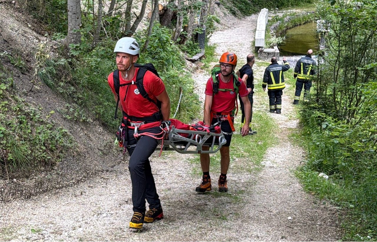 Zwei Bergretter tragen eine Trage, im Hintergrund stehen Feuerwehrleute auf einem Waldweg.