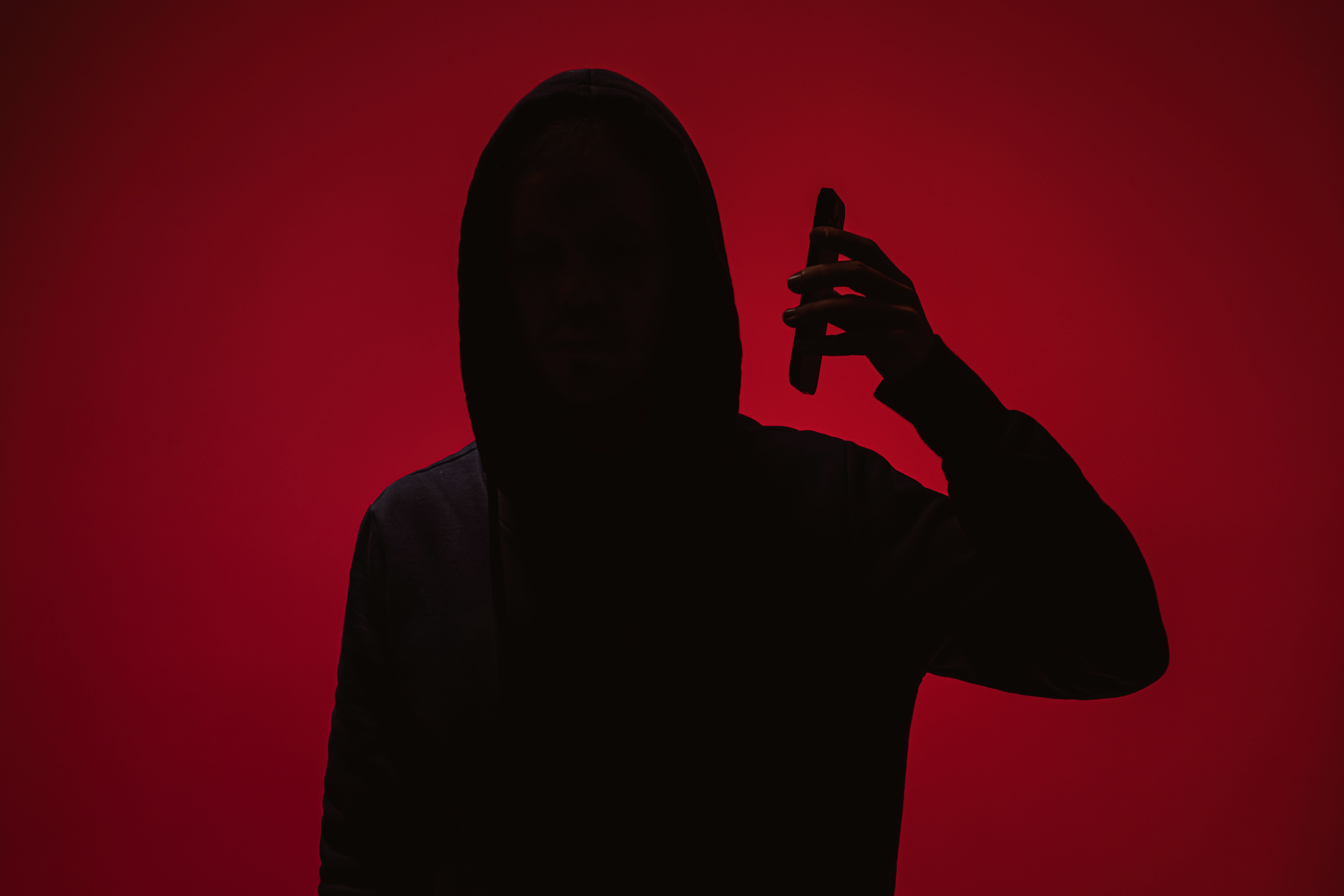 Silhouette einer Person mit Kapuze hält ein Handy vor rotem Hintergrund.