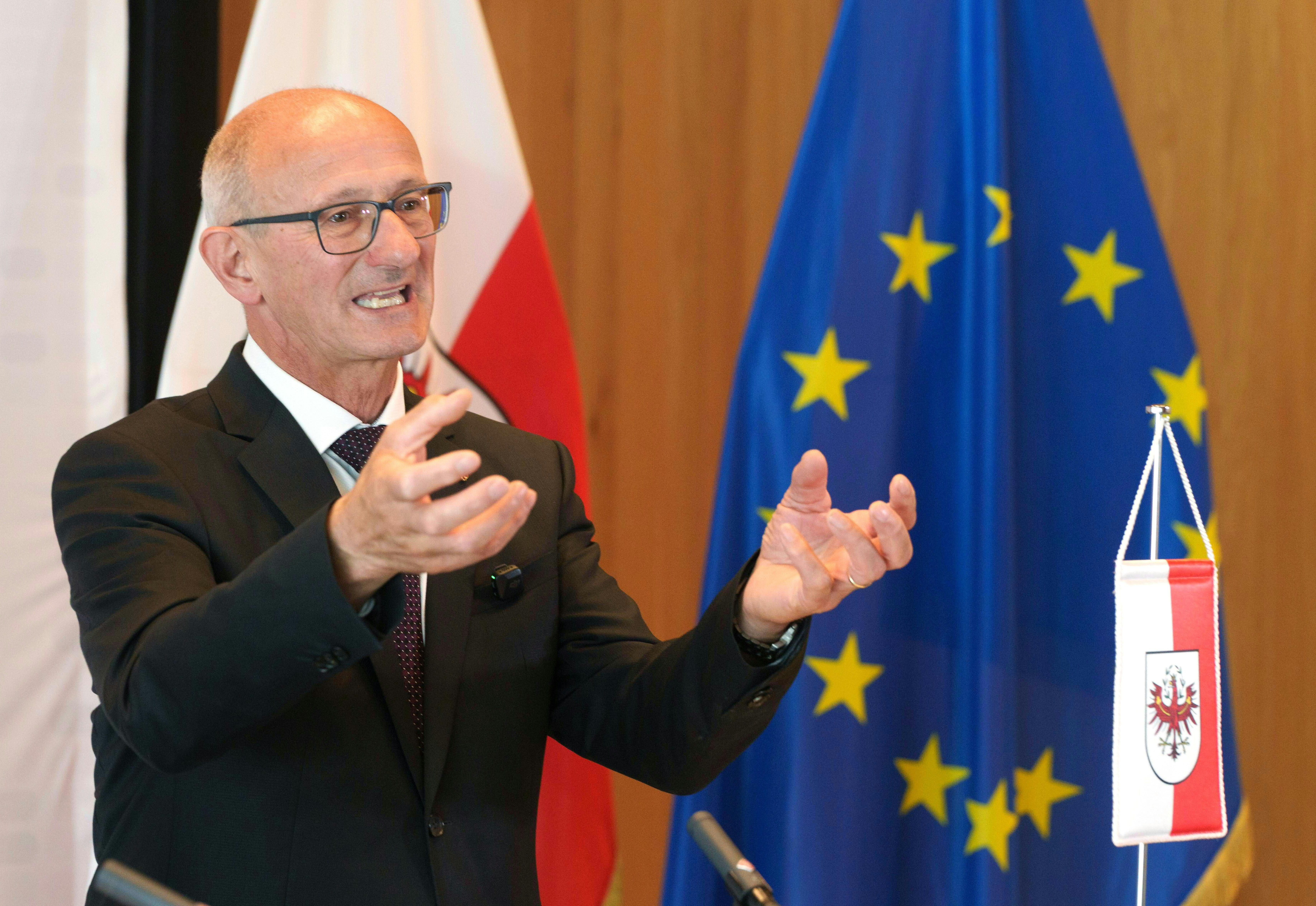 Ein Mann im Anzug spricht gestikulierend vor EU- und Tirol-Fahnen bei einer Pressekonferenz.