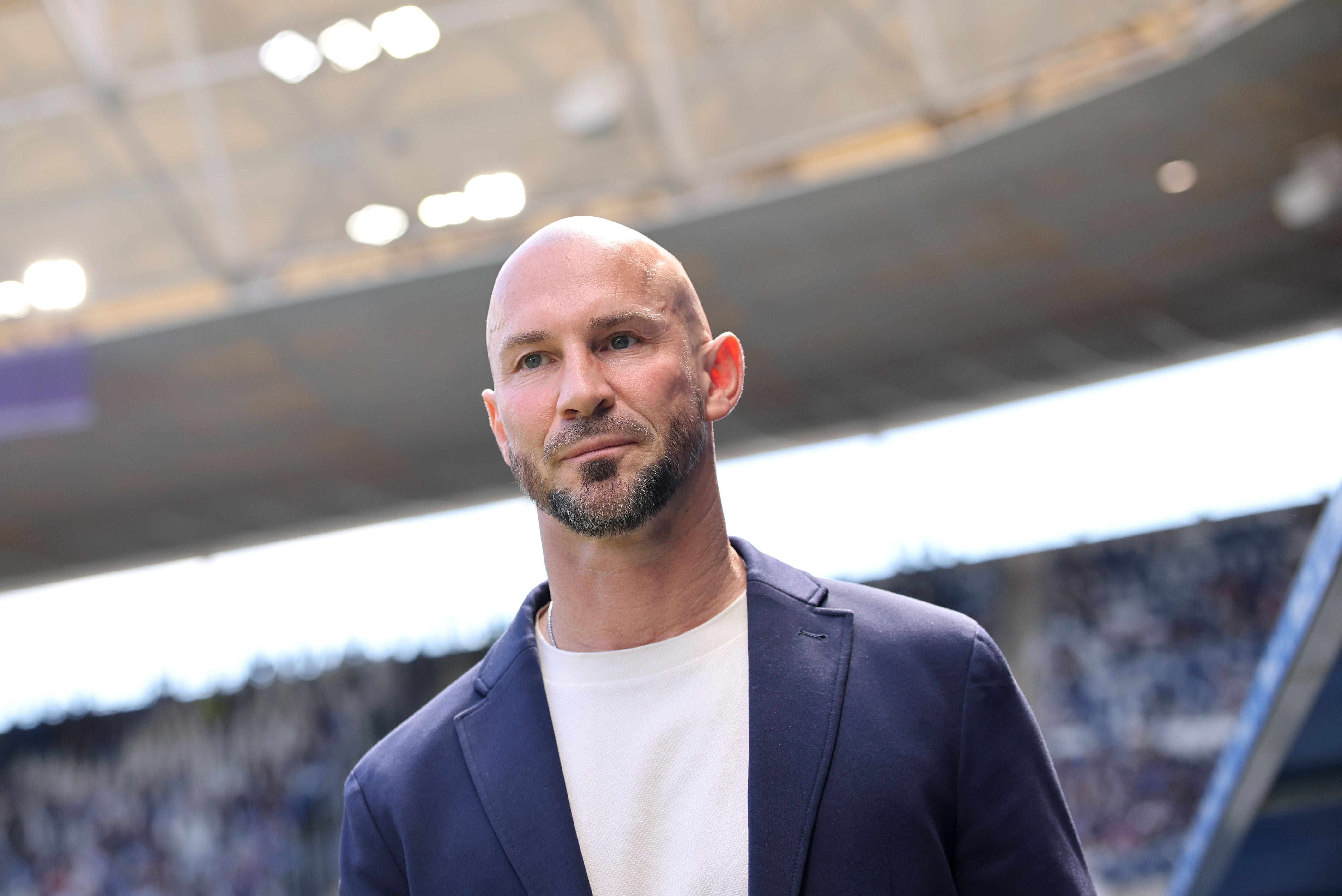 Ein Mann mit Bart und Glatze steht in einem Stadion und blickt nach vorn.