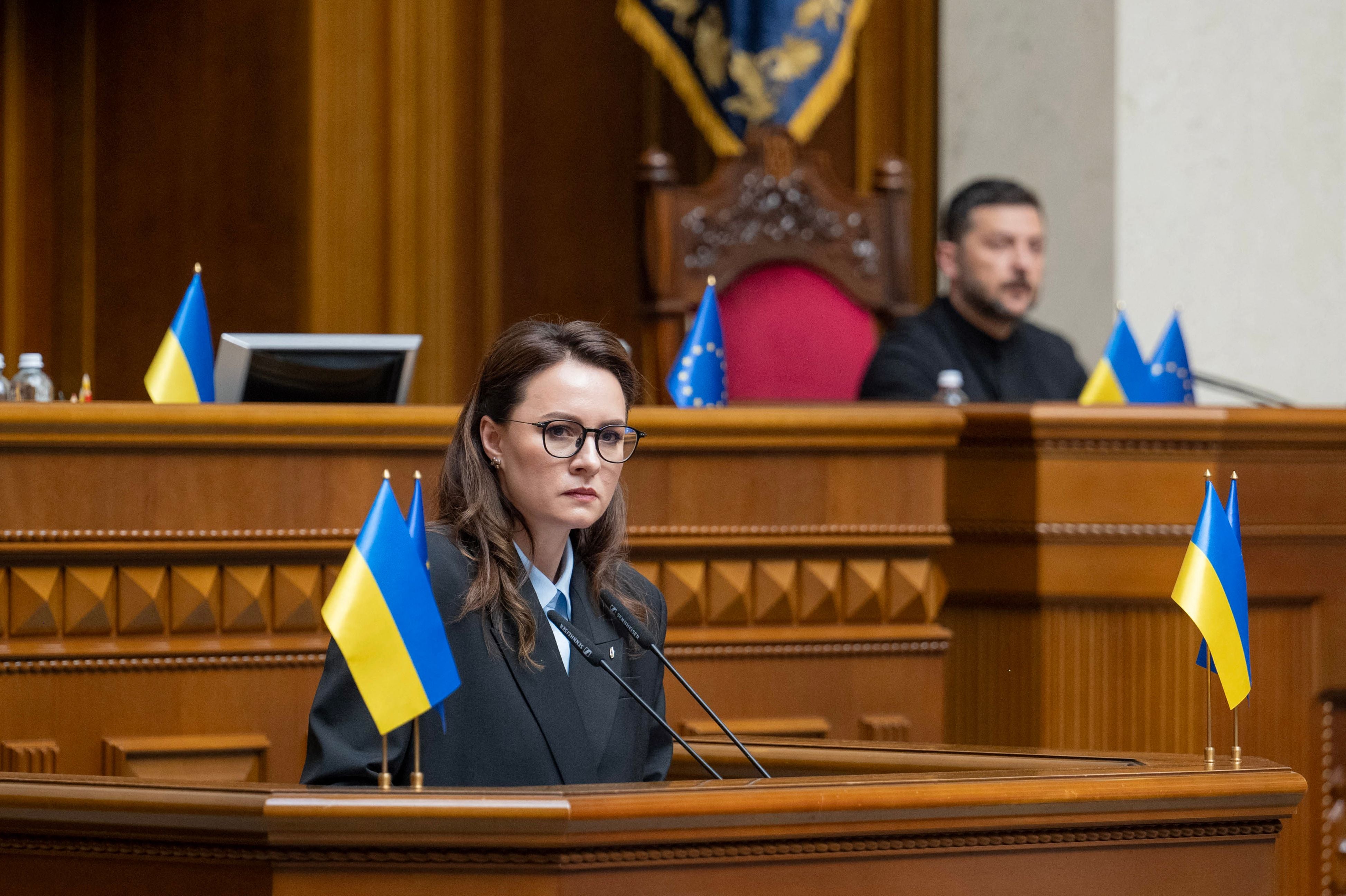 Eine Frau am Rednerpult im ukrainischen Parlament, flankiert von ukrainischen Flaggen.